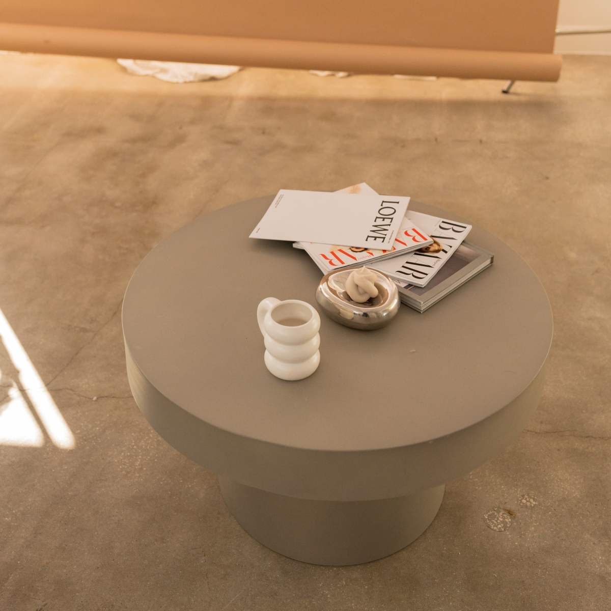 A round, gray coffee table sits on a concrete floor with a stack of magazines and a small dish on top, alongside a white mug.