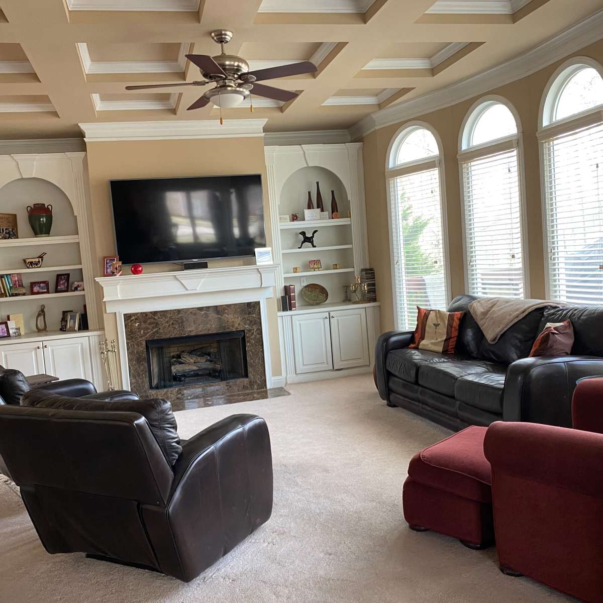 A living room features a coffered ceiling, a television mounted above a fireplace, and three seating arrangements, including a red chair, in front of large windows.