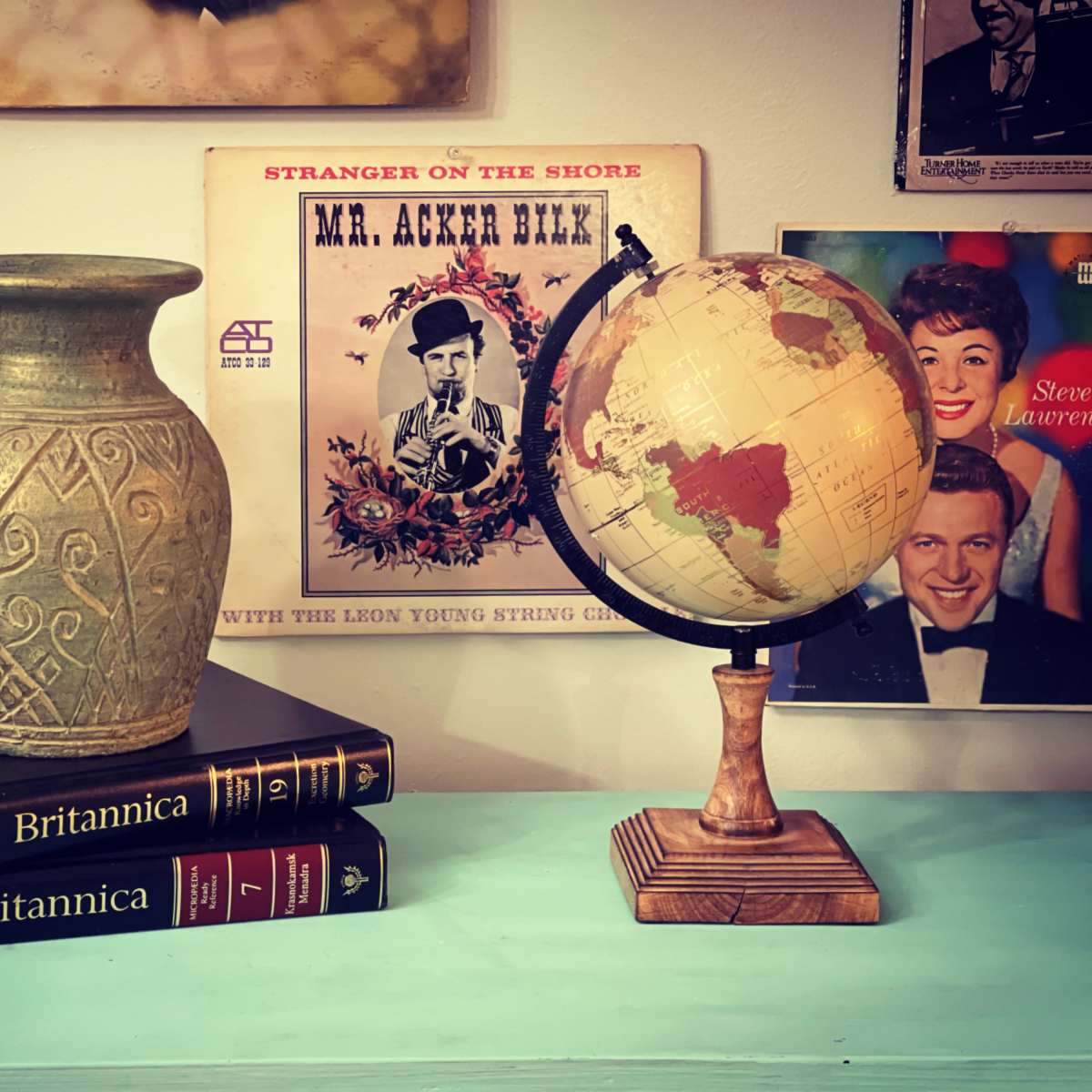 A decorative vase sits atop two volumes of the "Britannica" encyclopedia, alongside a vintage globe on a wooden stand, with framed records on the wall in the background.