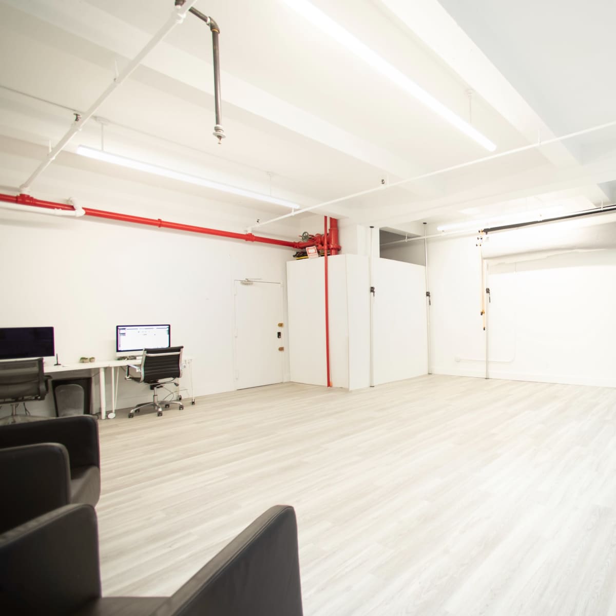 A spacious, well-lit room with light wood flooring and minimal furniture, featuring a large white wall suitable for presentations or photography.