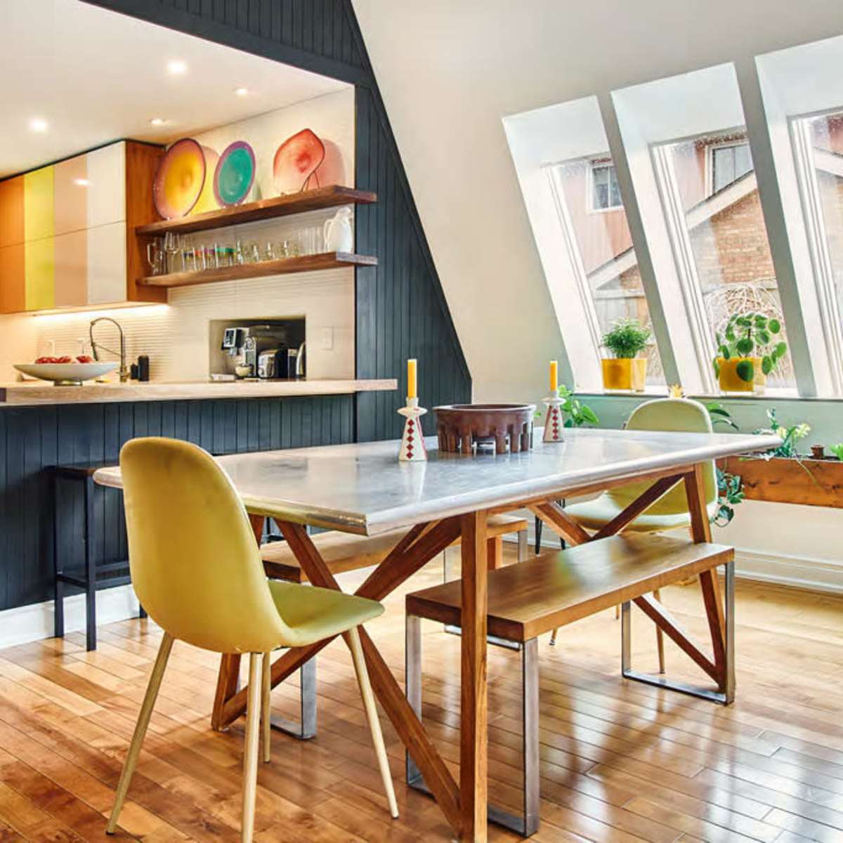 A modern kitchen-dining area features a marble table surrounded by wooden benches and yellow chairs, with large angled windows and colorful dishware displayed on the wall.