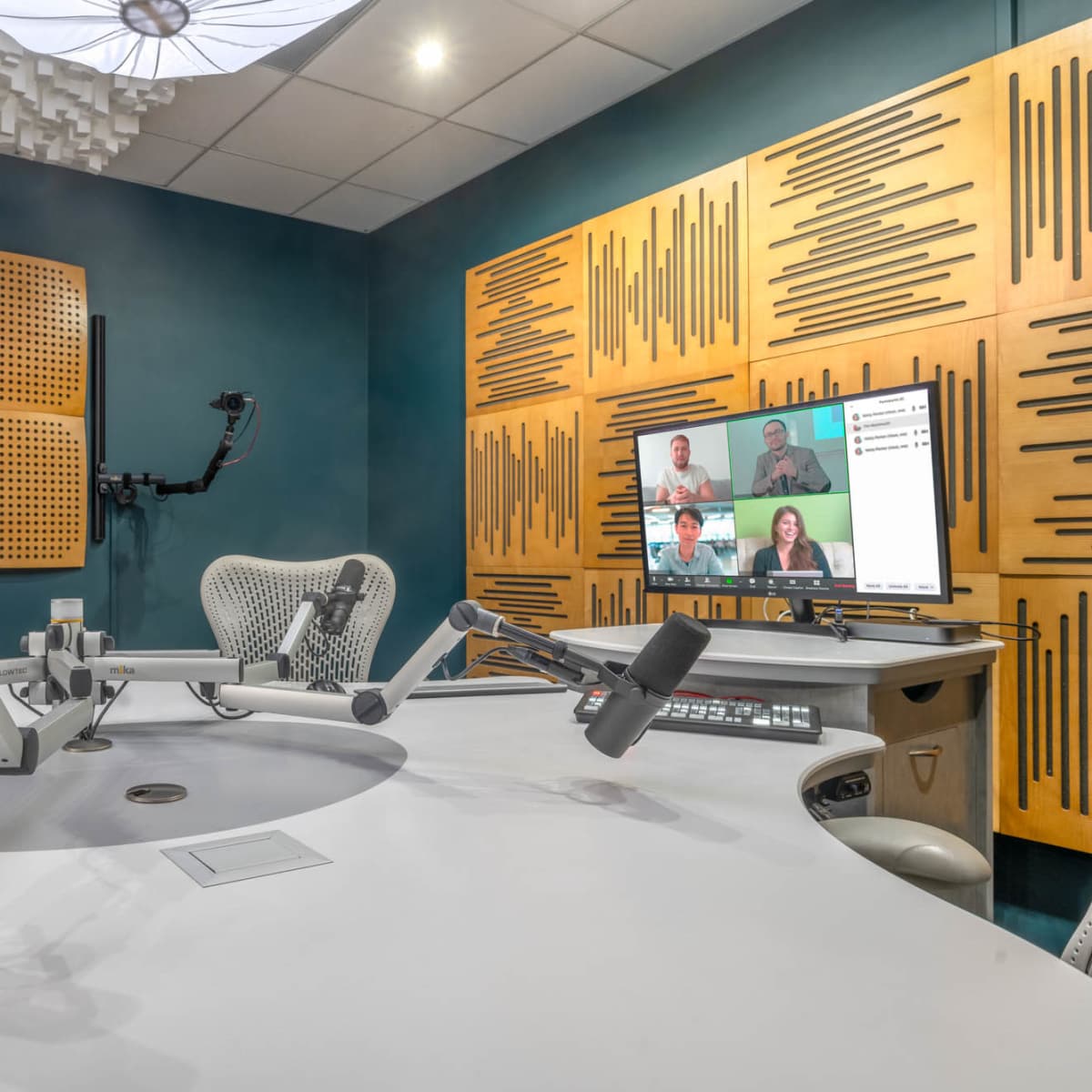 A modern recording studio with a round white table, multiple microphones, soundproof walls, and a monitor displaying video participants.
