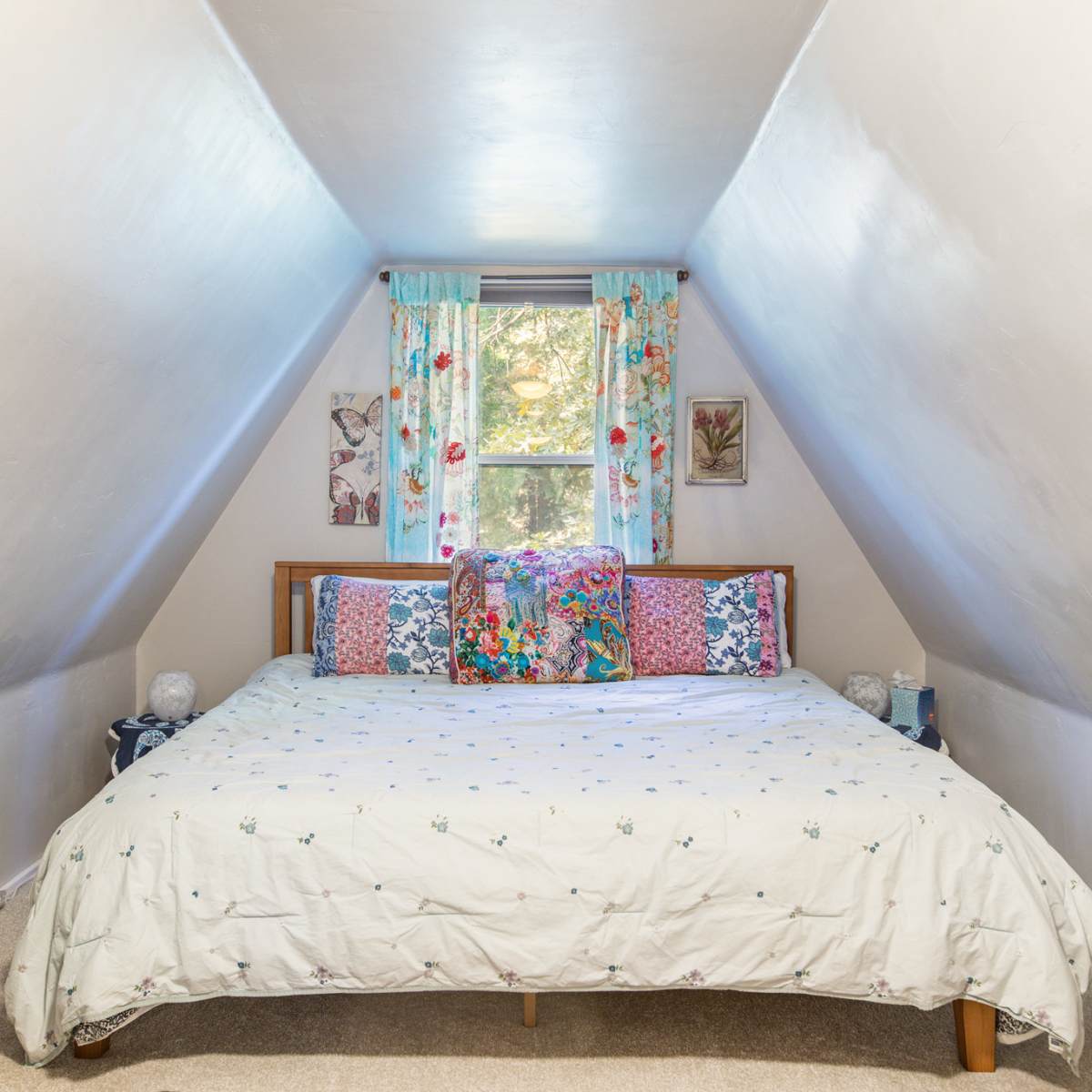 A cozy attic bedroom with a double bed, patterned pillows, and a window adorned with floral curtains.