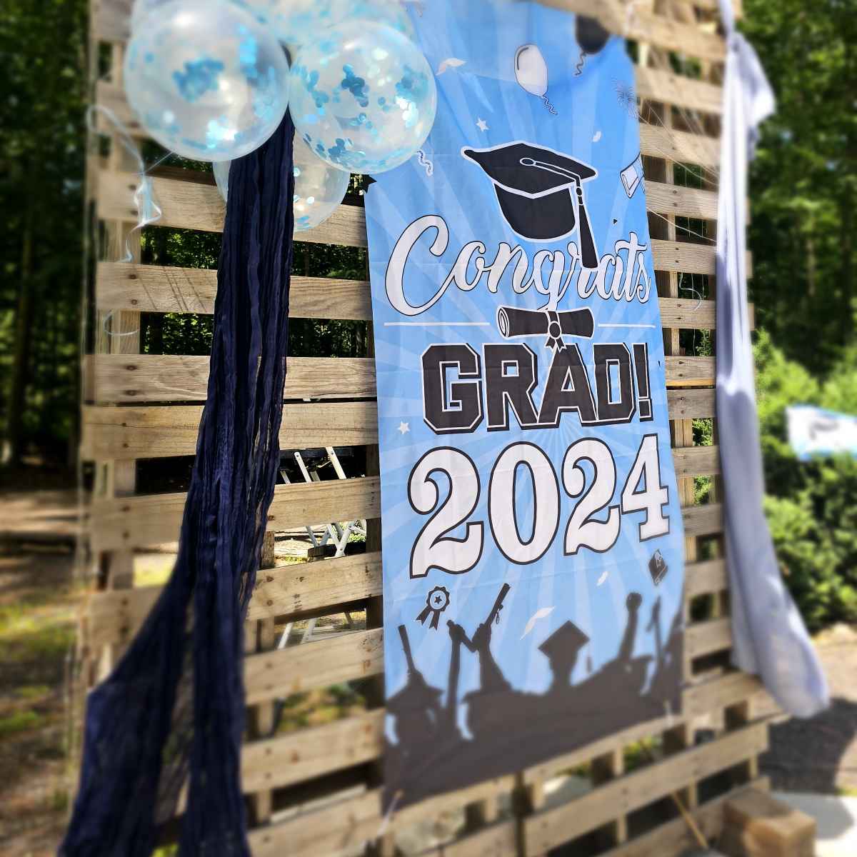 A banner reading "Congrats GRAD! 2024" is displayed alongside blue and white balloons in a decorated outdoor setting.