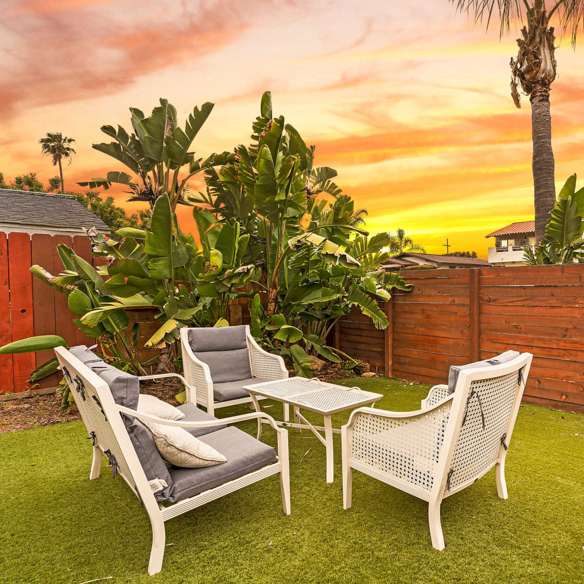 A set of white outdoor furniture is arranged on green grass in a backyard surrounded by tall plants and a wooden fence, with a vibrant sunset sky in the background.