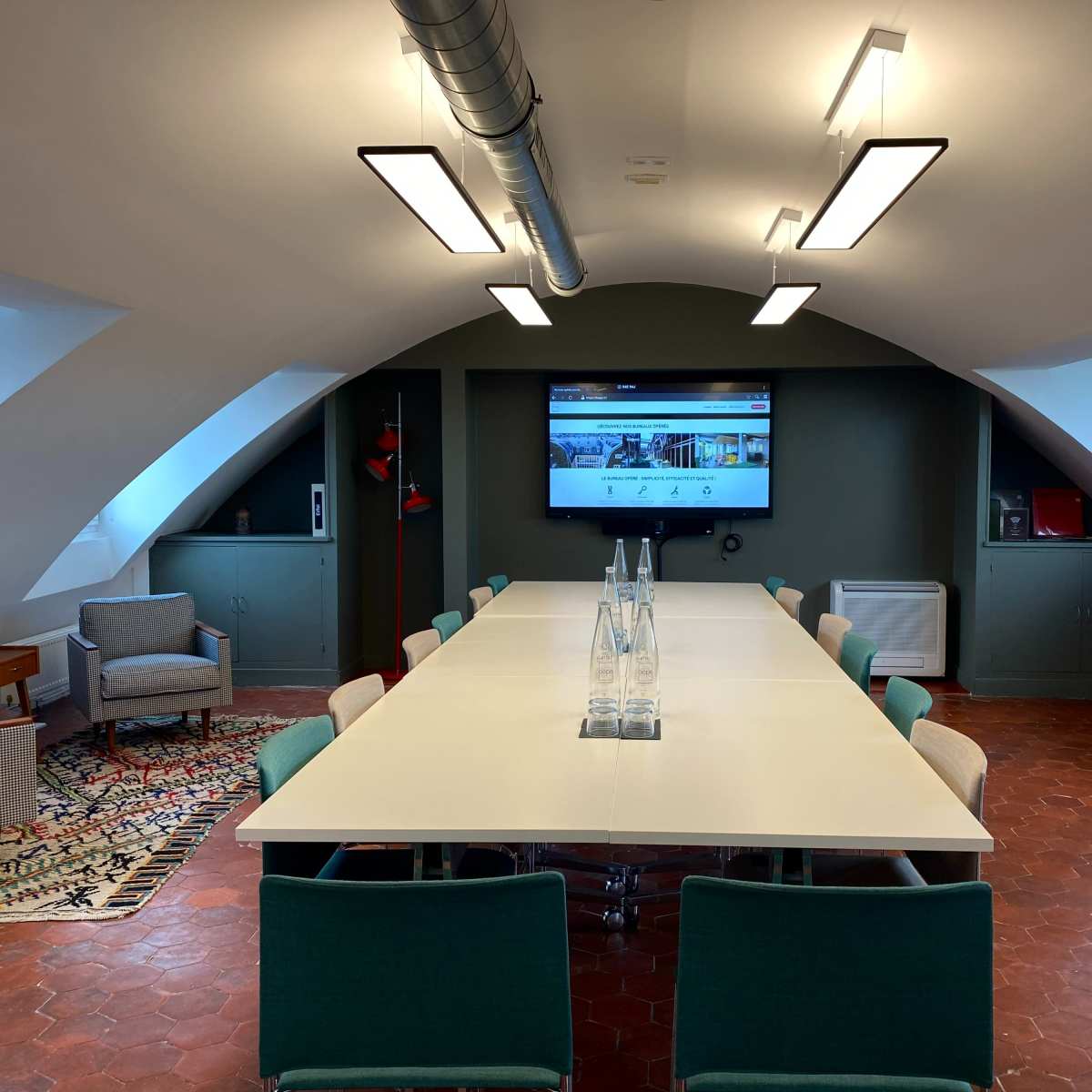 A spacious conference room features a long table surrounded by chairs, with a large screen displaying content on the wall and natural light coming through the windows.