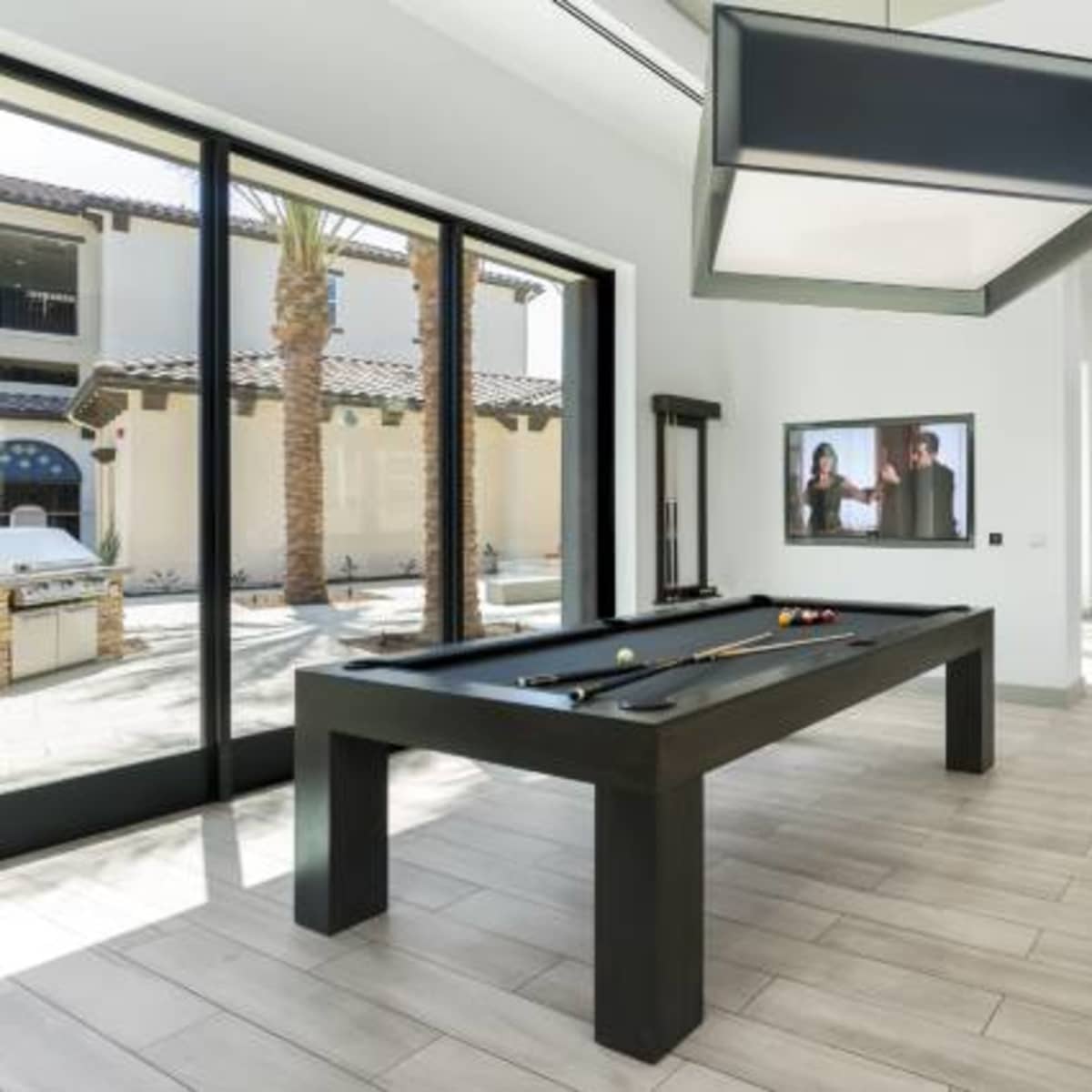 A game room features a black pool table and large windows overlooking a courtyard with palm trees.