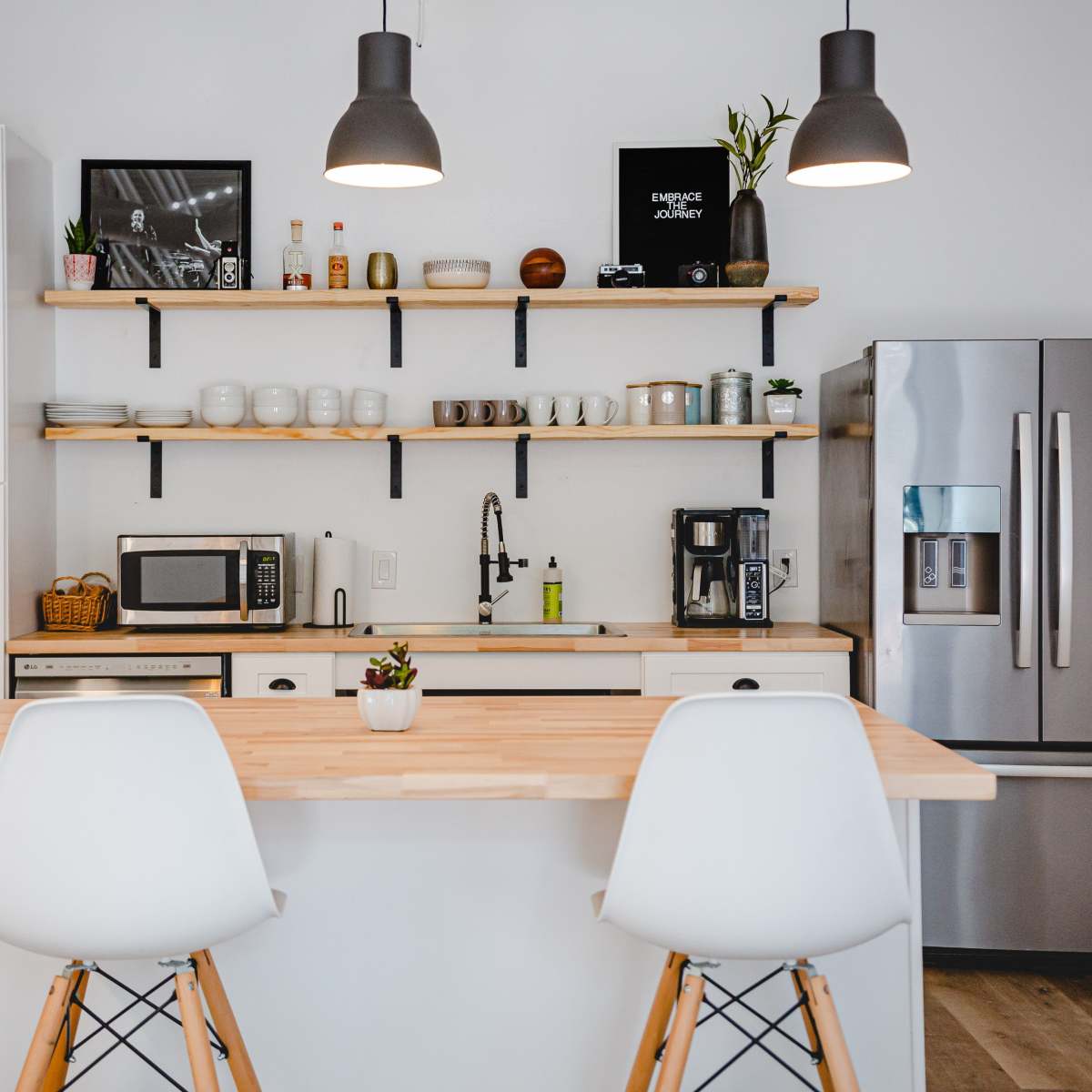 The kitchen features sleek white cabinets, a wooden countertop with two stools, open shelving displaying dishware, and modern appliances including a refrigerator and microwave.