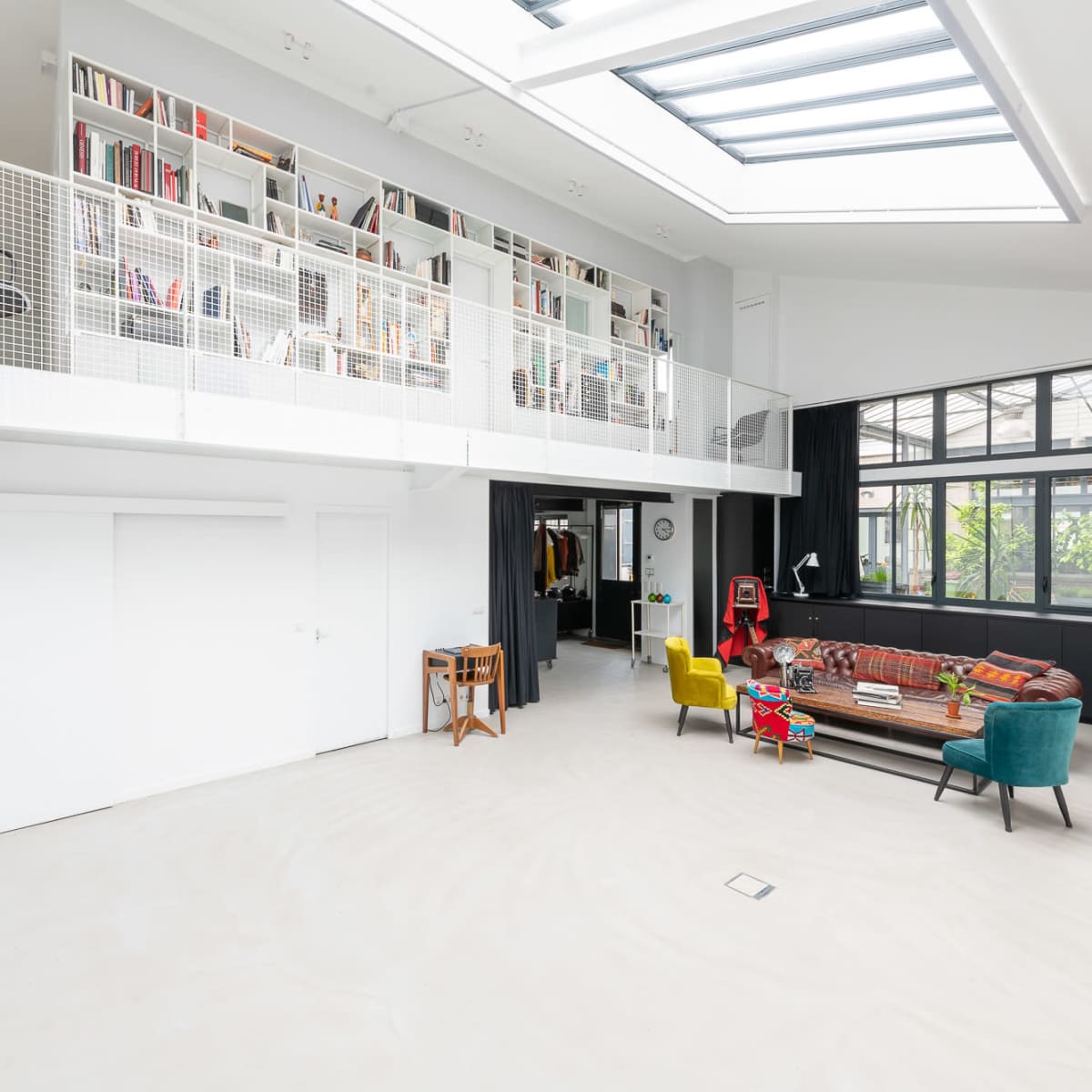 A modern, open-concept living space with a high ceiling, featuring a mezzanine library, large windows, and a colorful furniture arrangement.