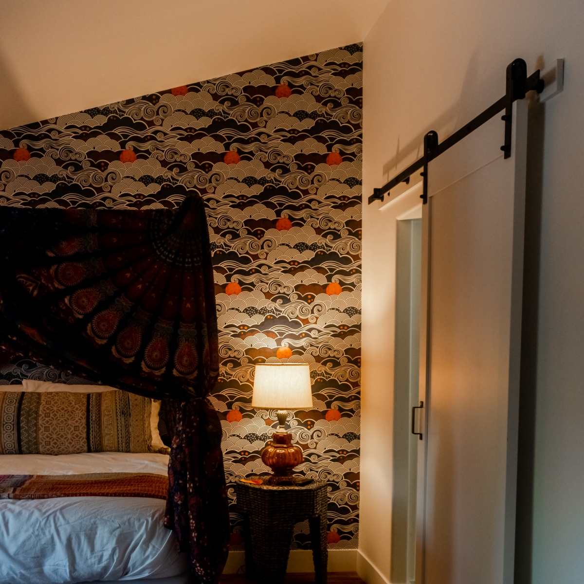 A bedroom with a patterned wallpaper in the background, a bed with a canopy, a lamp on a side table, and a sliding door.