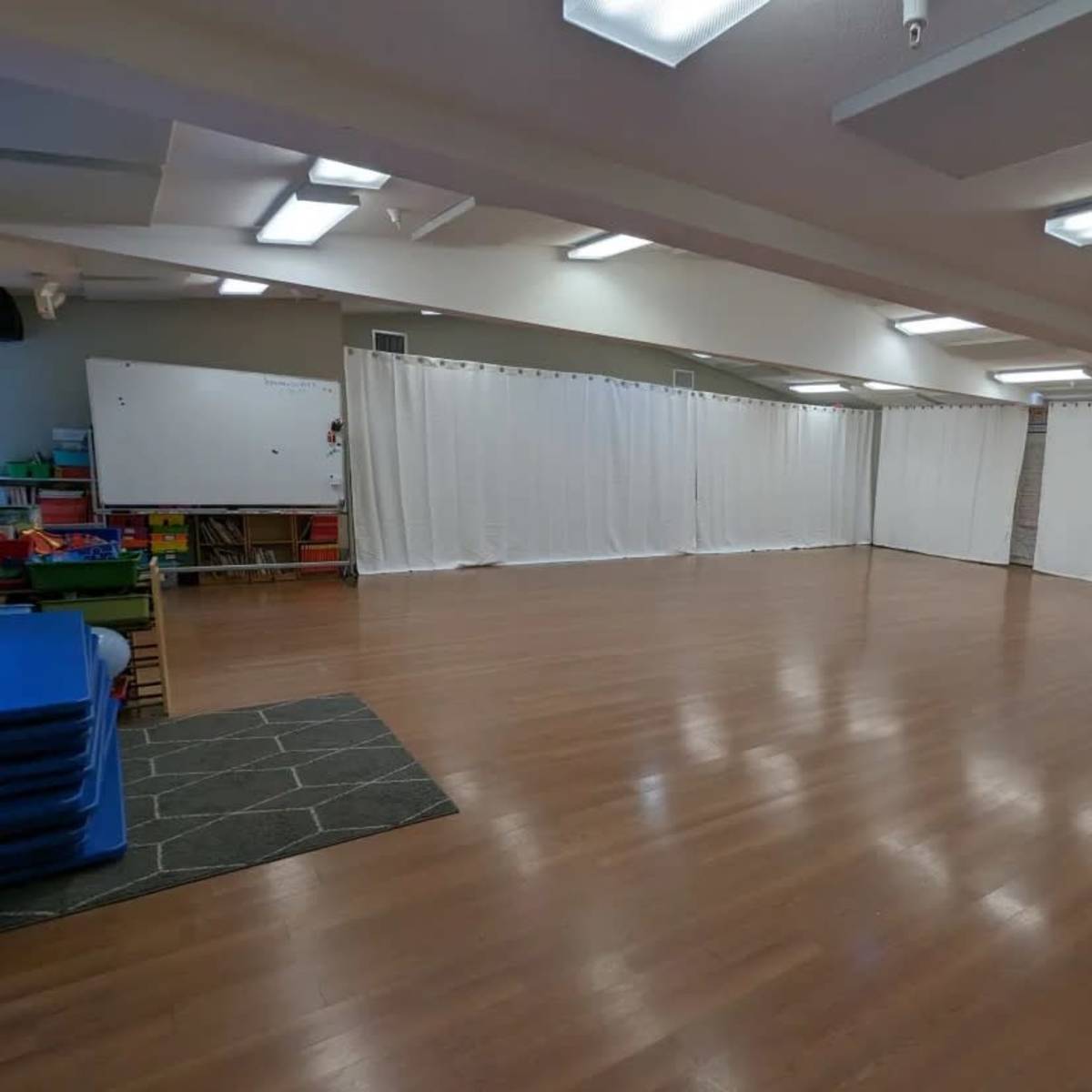 A spacious indoor room with wooden floors, white curtains dividing the space, and neatly stacked blue mats in one corner.