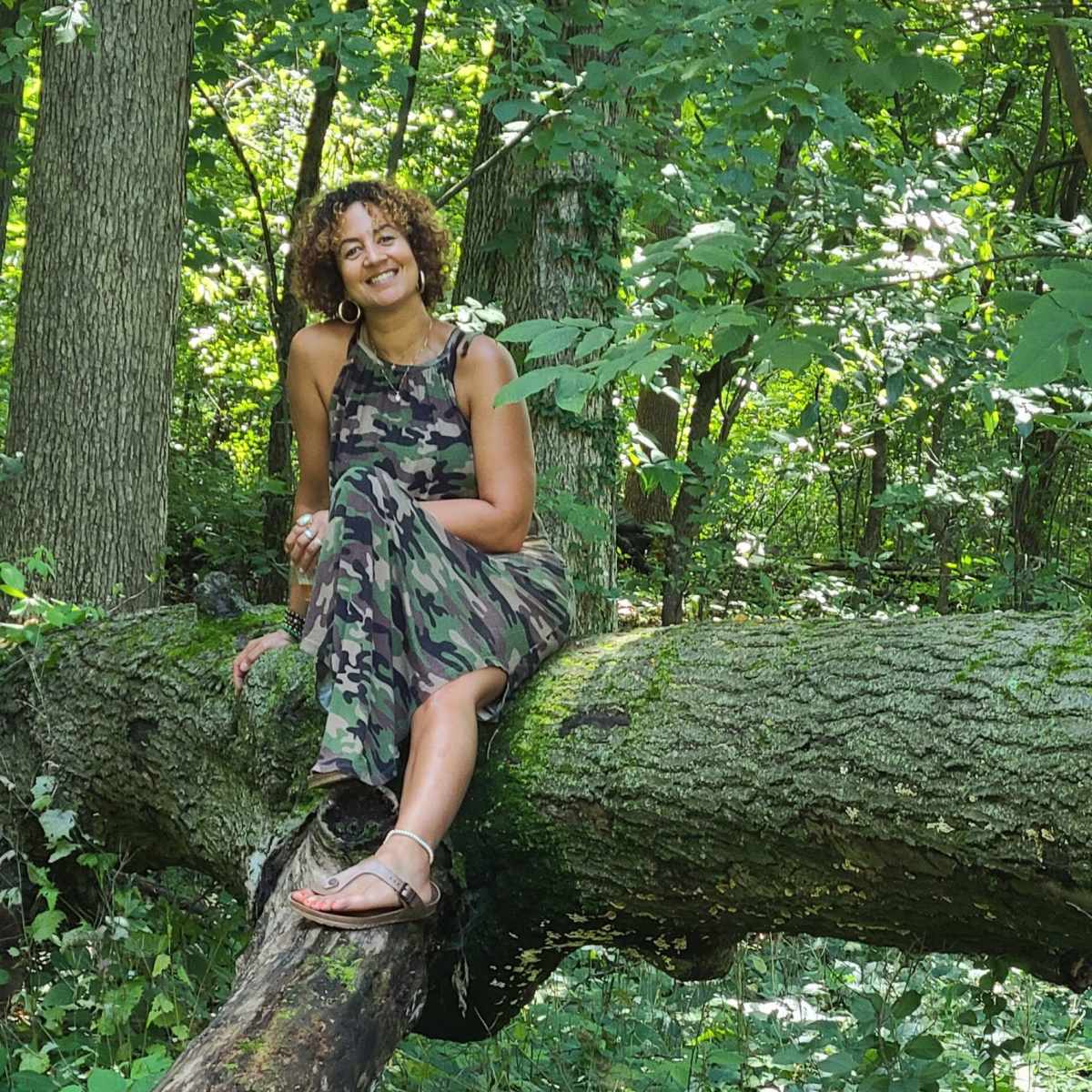 A woman sits on a fallen tree trunk in a wooded area, surrounded by greenery.