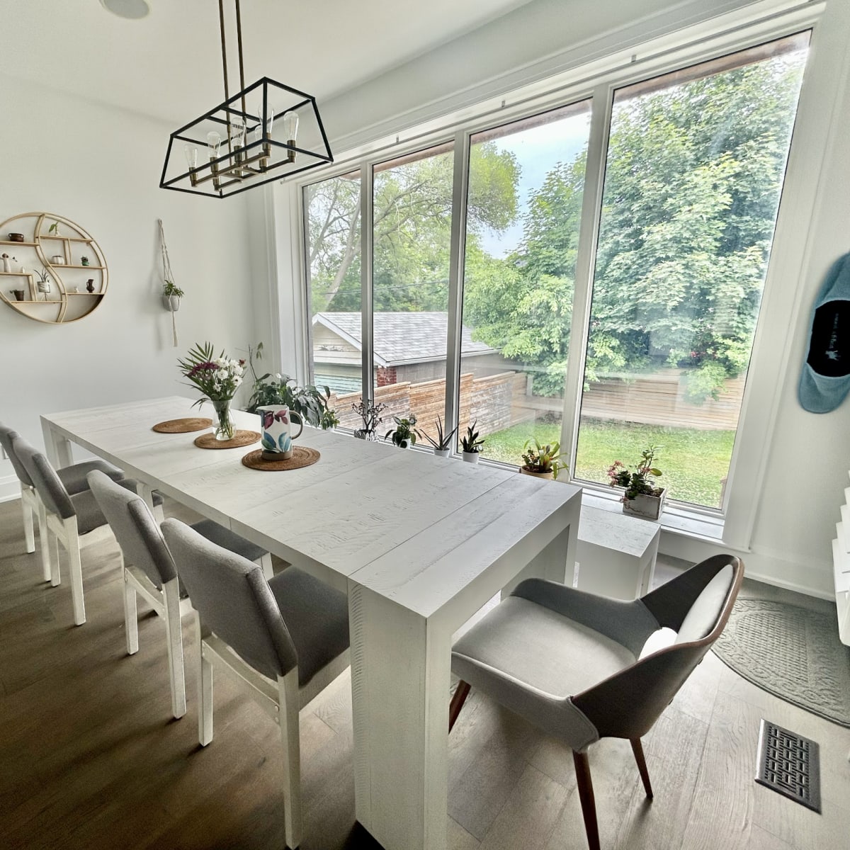 A bright dining area features a long white table with gray chairs, surrounded by large windows offering a view of a green outdoor space.