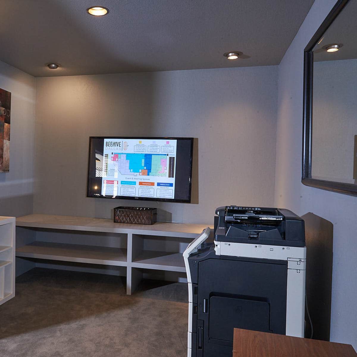 A small office space featuring a wall-mounted TV, a printer, a storage unit with cubicles, and a mirror.