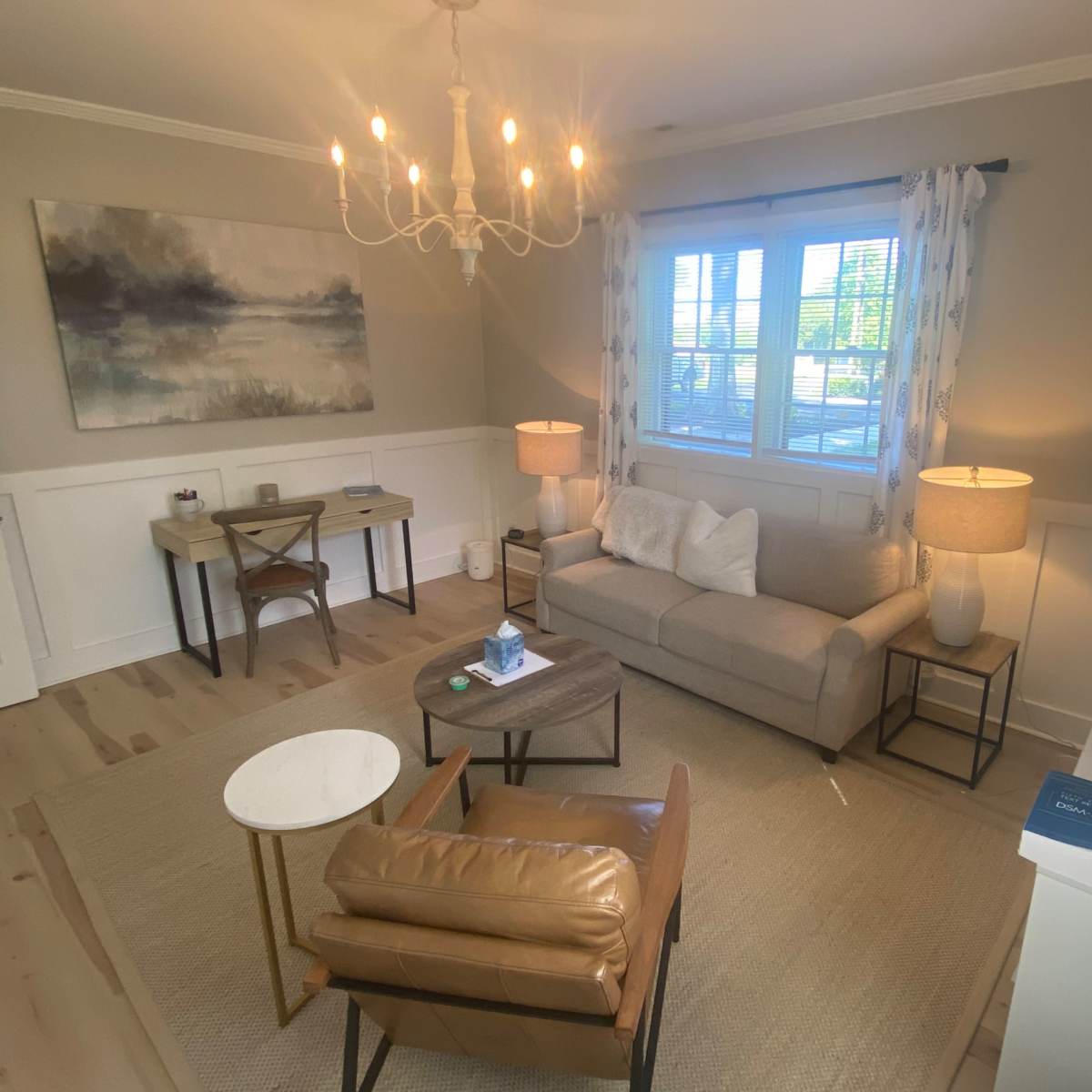 A cozy living room with a light-colored sofa, two lamps, a coffee table, and a workspace in the corner.