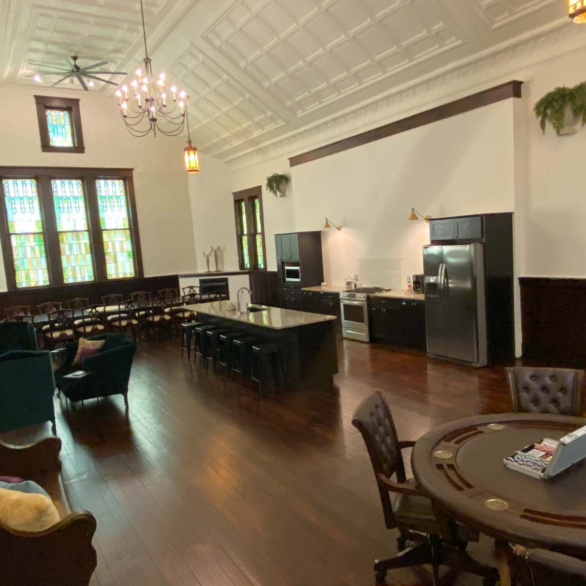 A spacious room with a high ceiling, featuring dining tables on one side and a kitchen area on the other, along with stained glass windows and wooden floors.