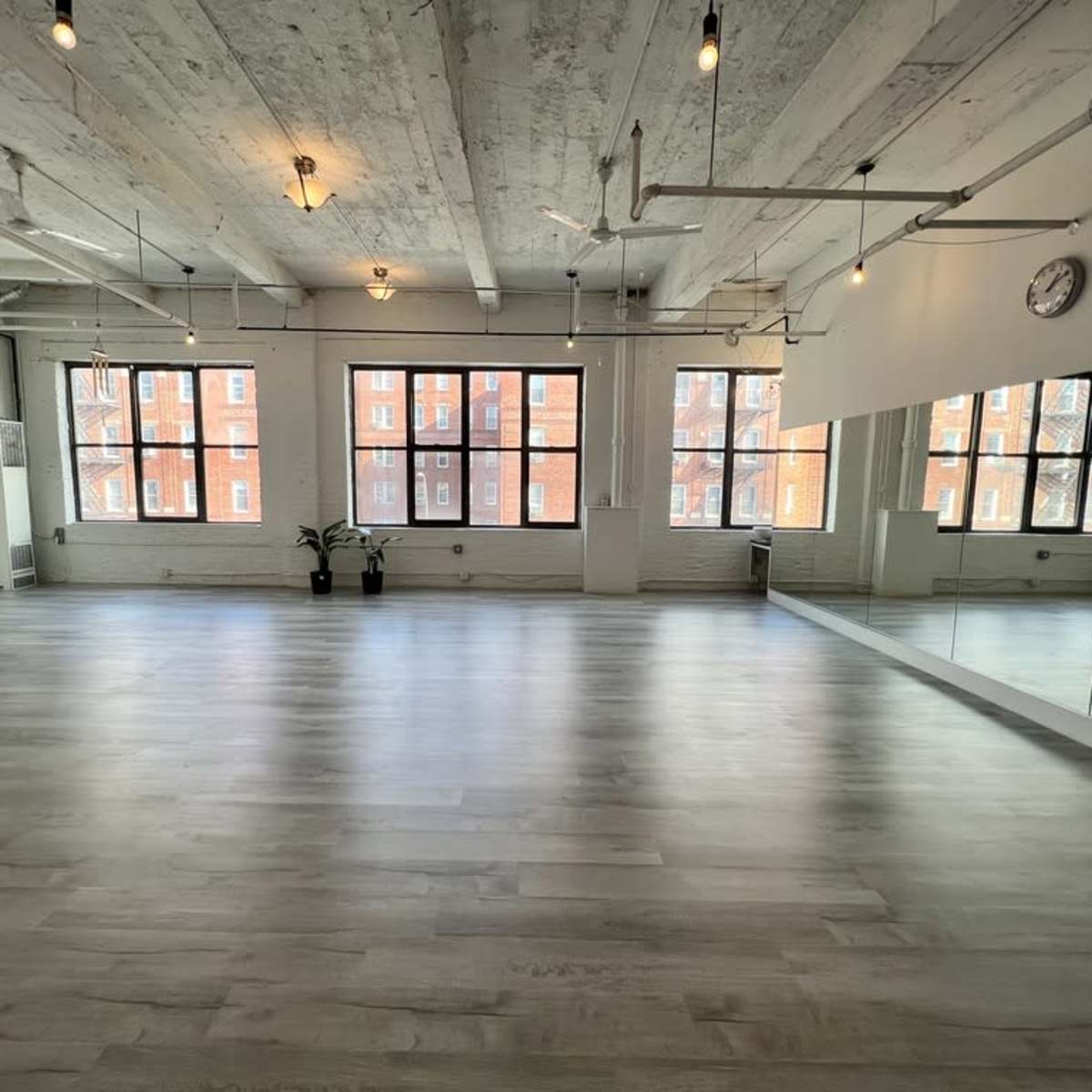 A spacious, well-lit studio with large windows, wood-like flooring, and a mirror along one wall.