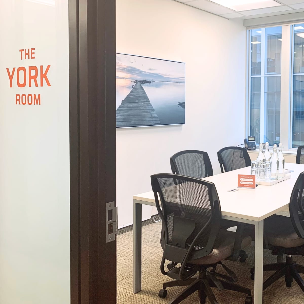 A conference room with a large white table surrounded by six chairs and a large screen displaying a waterfront image.