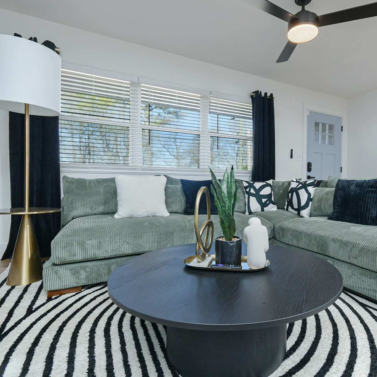 The living room features a green sectional sofa, a round black coffee table with decorative items, and large windows allowing natural light to enter the space.