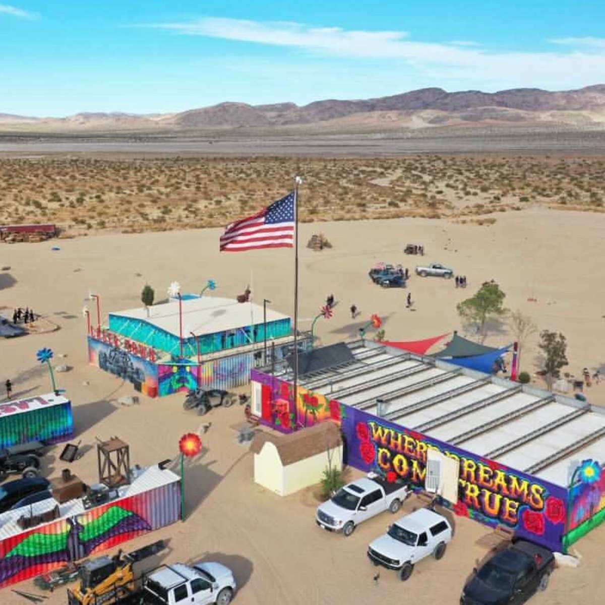 An aerial view shows a vibrant gathering in a desert landscape, featuring colorful structures, vehicles, and an American flag.