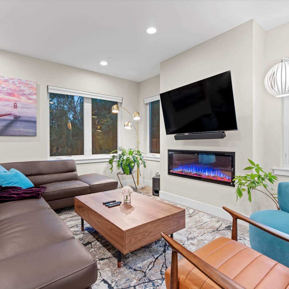 The living room features a sectional sofa, a modern coffee table, a mounted TV, and a wall-mounted electric fireplace, with large windows providing natural light.