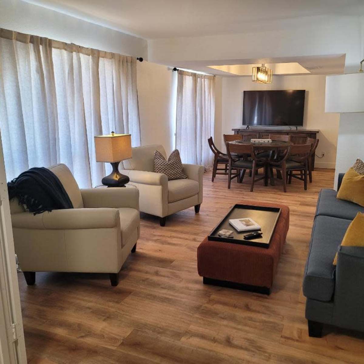 A modern living room featuring two armchairs, a sofa, a coffee table, a dining area in the background, and large windows with sheer curtains.