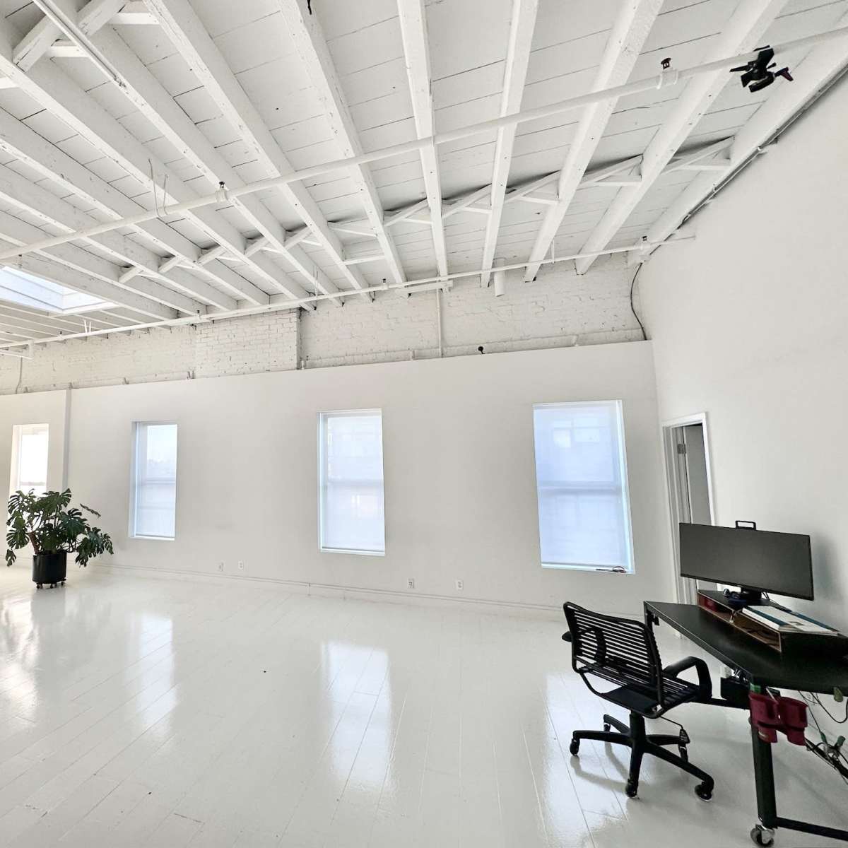 A spacious, modern room with white walls, a white ceiling, several windows, and a black desk with a computer.