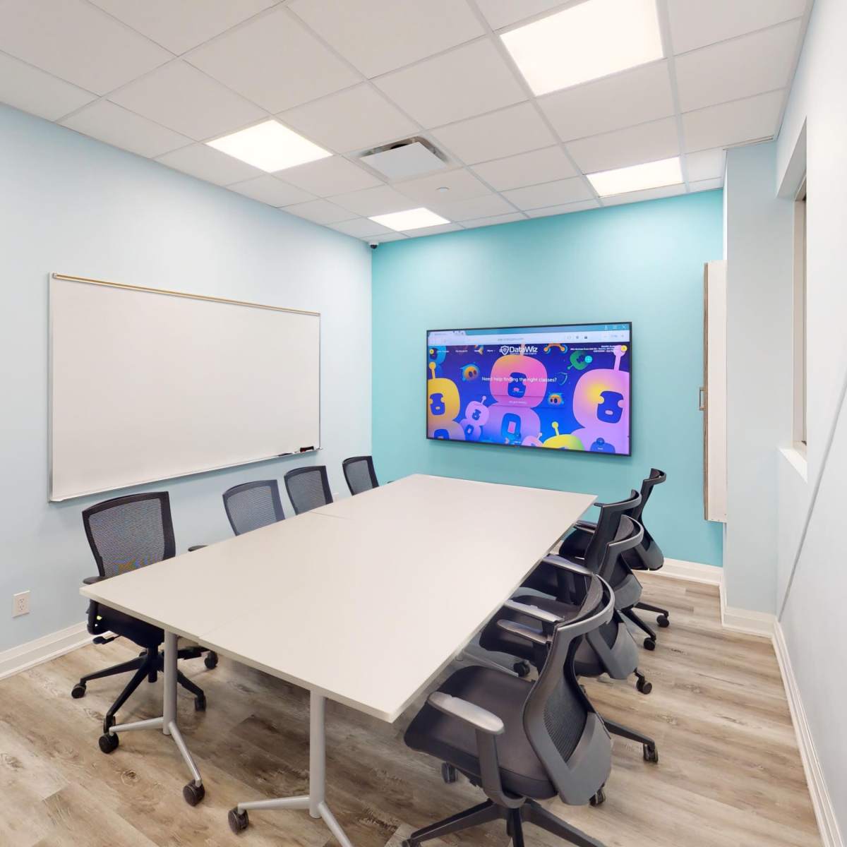 A meeting room features a long white table surrounded by ergonomic chairs, a whiteboard, and a large screen displaying colorful graphics on a light blue wall.