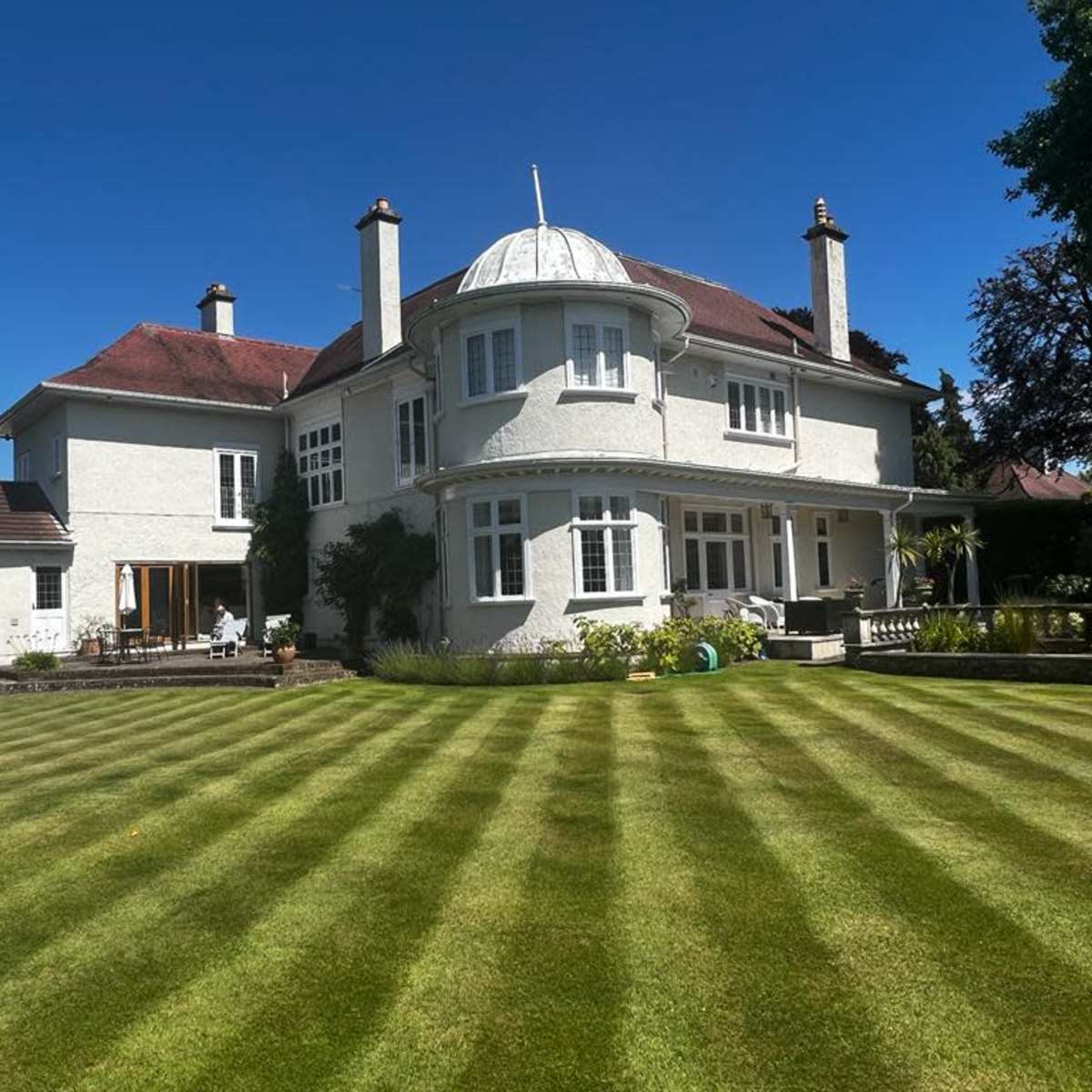 A large, white mansion with a rounded turret and well-maintained lawn features neatly mowed stripes under a clear blue sky.