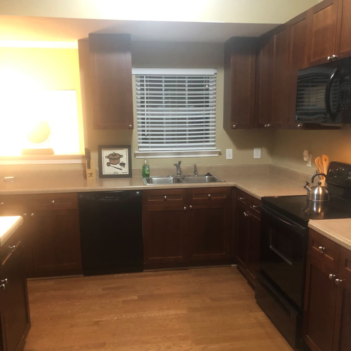 A kitchen with wooden cabinets, a black dishwasher, and an oven, featuring a window with blinds and a counter with kitchen utensils.