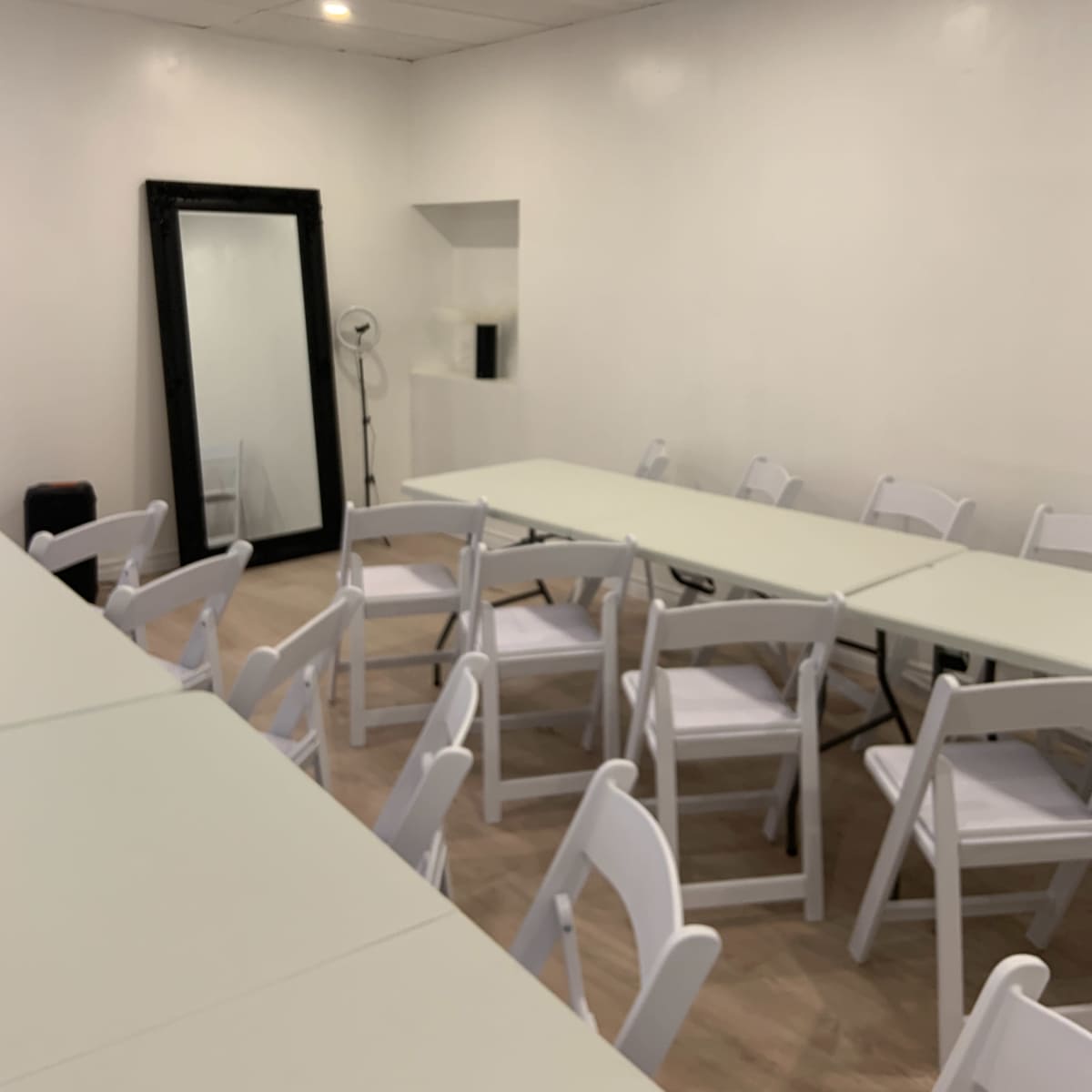 A sparsely furnished room with several tables arranged in a row and white folding chairs set around them, along with a large mirror on one wall.