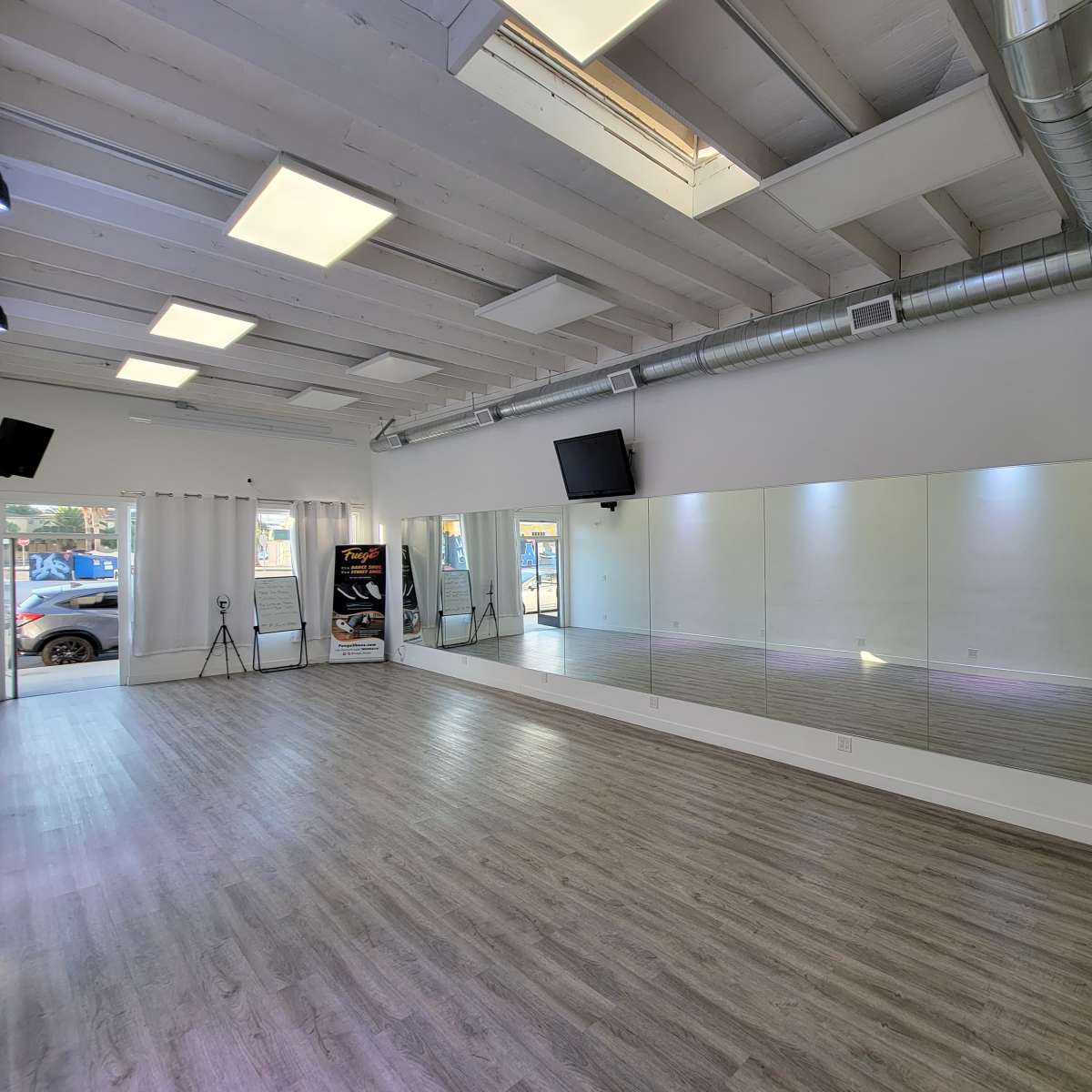 A spacious dance studio with wooden flooring, large mirrors on one wall, and natural light streaming in through a window.