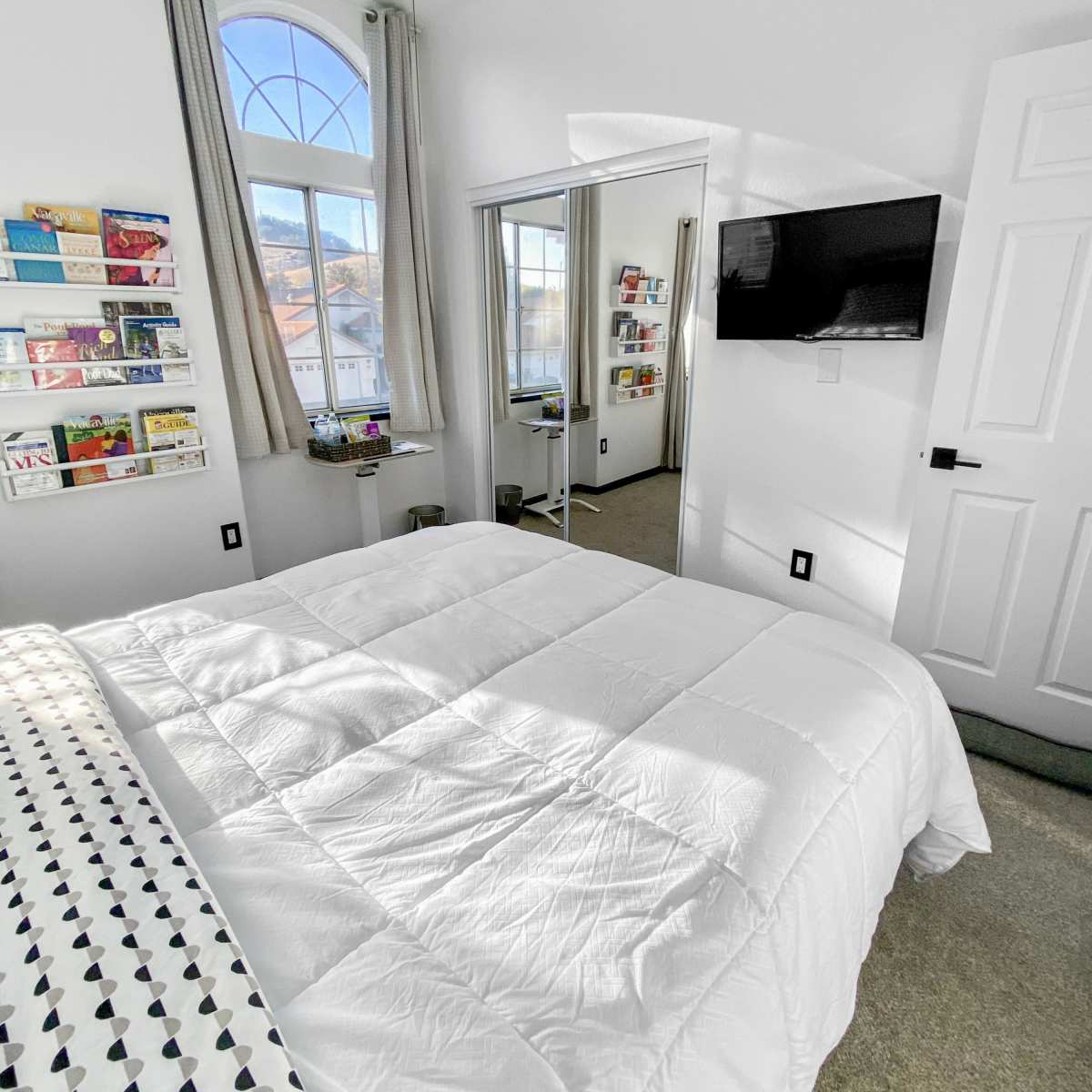 A neatly arranged bedroom features a white bedspread, a wall-mounted TV, and a large window with curtains.