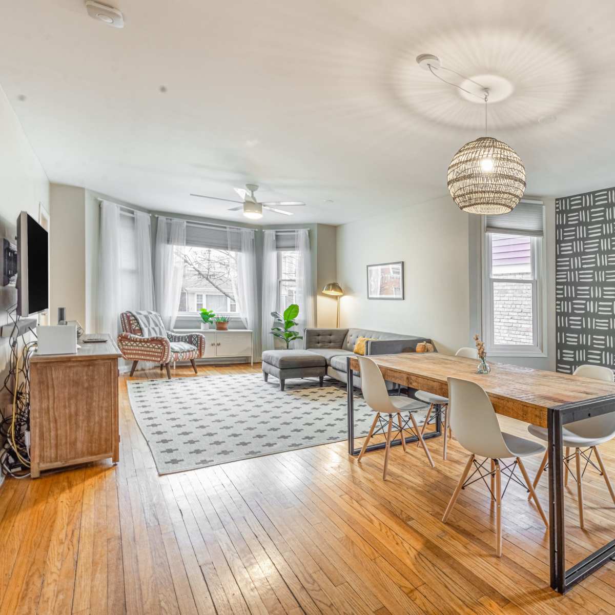 A spacious living room and dining area featuring a wooden table, gray chairs, a patterned rug, and large windows with natural light.