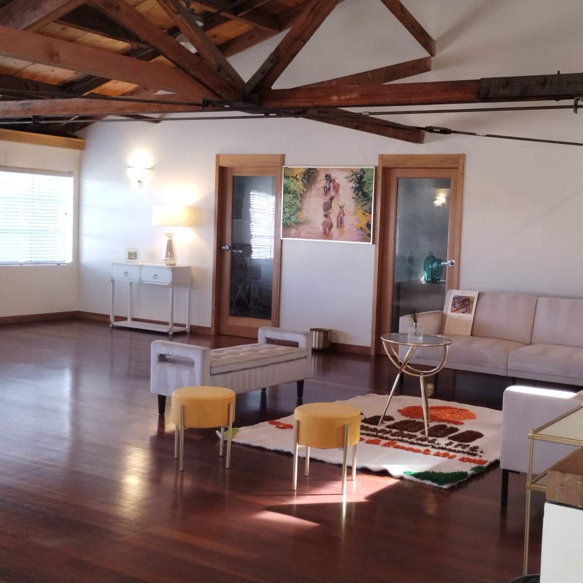 A spacious room with wooden beams, a light-colored sofa, a coffee table, and a decorative rug, featuring a side table and large windows.