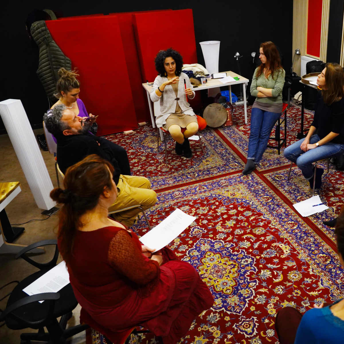 A group of people is seated in a circle on a patterned rug in a studio, engaging in a discussion led by a woman in the center.
