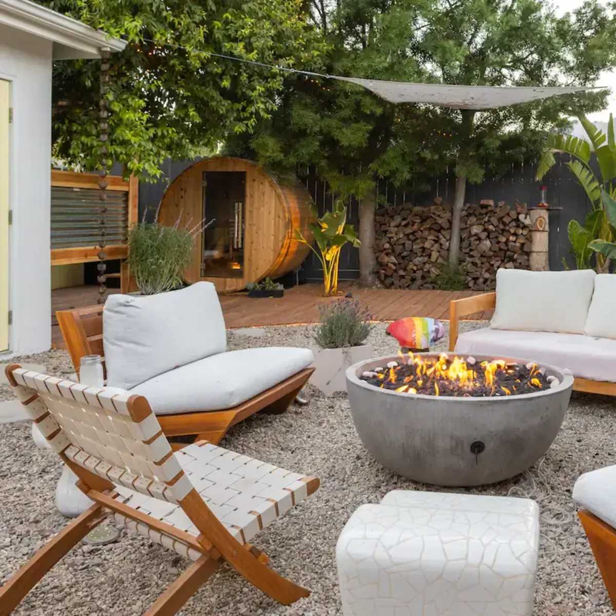 A circular fire pit surrounded by wooden lounge chairs and a stone table is set in a gravel patio area, with greenery and stacked firewood in the background.