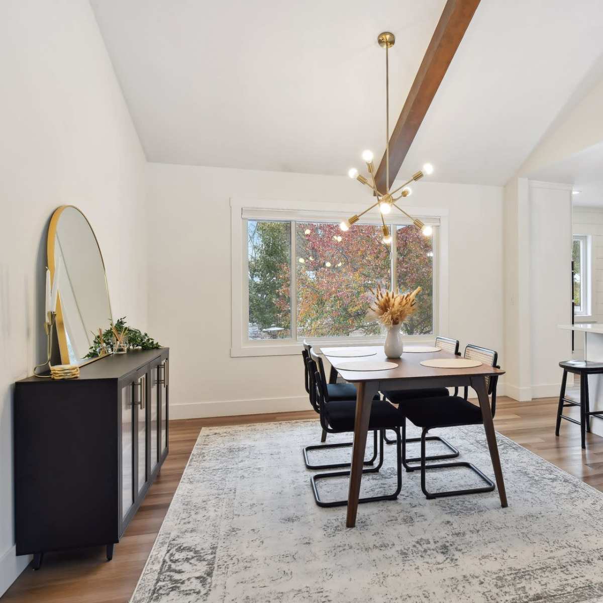 A modern dining area featuring a wooden table with black chairs, a sideboard, and a large window overlooking colorful trees.