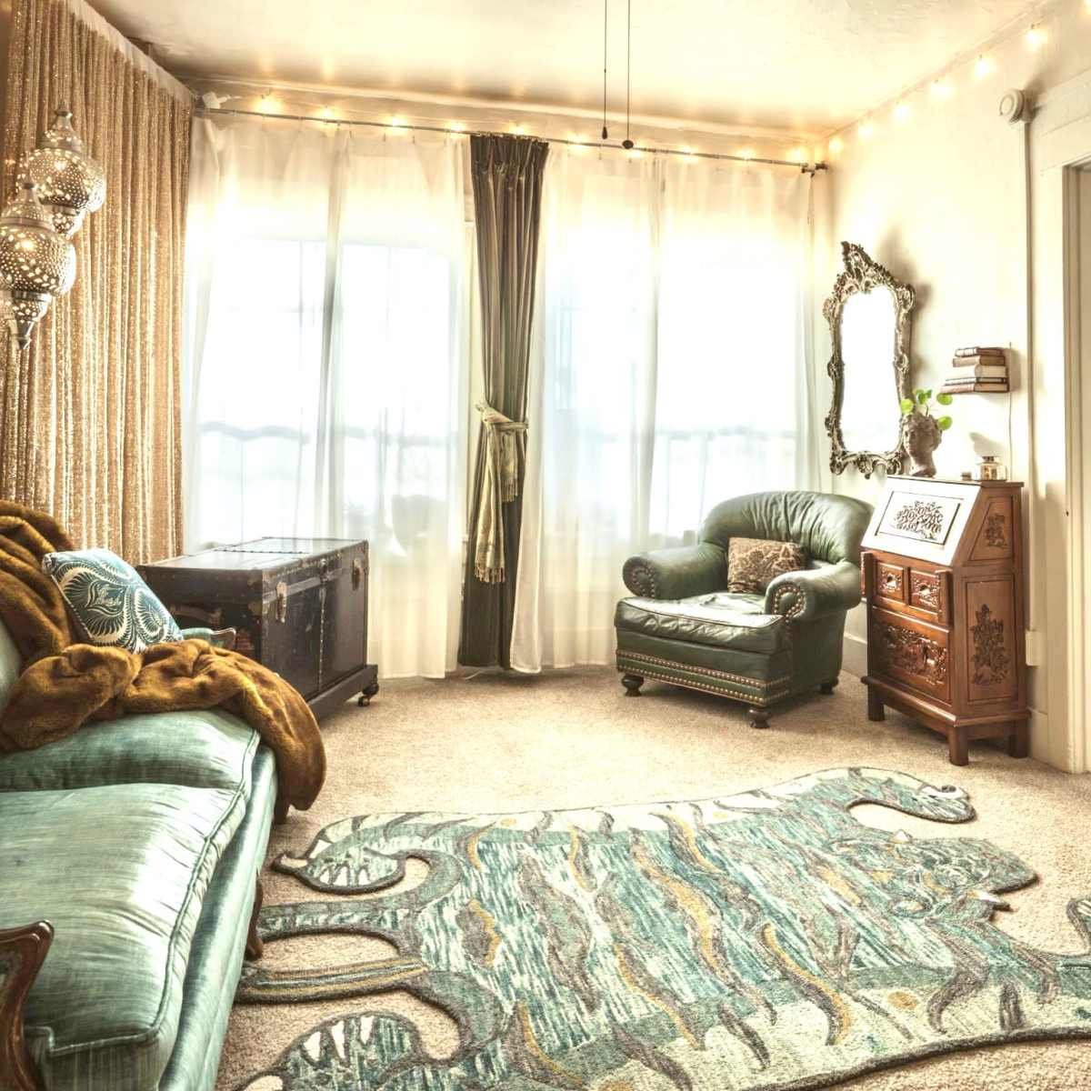 A cozy living room featuring a green sofa, a matching armchair, a decorative area rug, and large windows draped with sheer curtains.