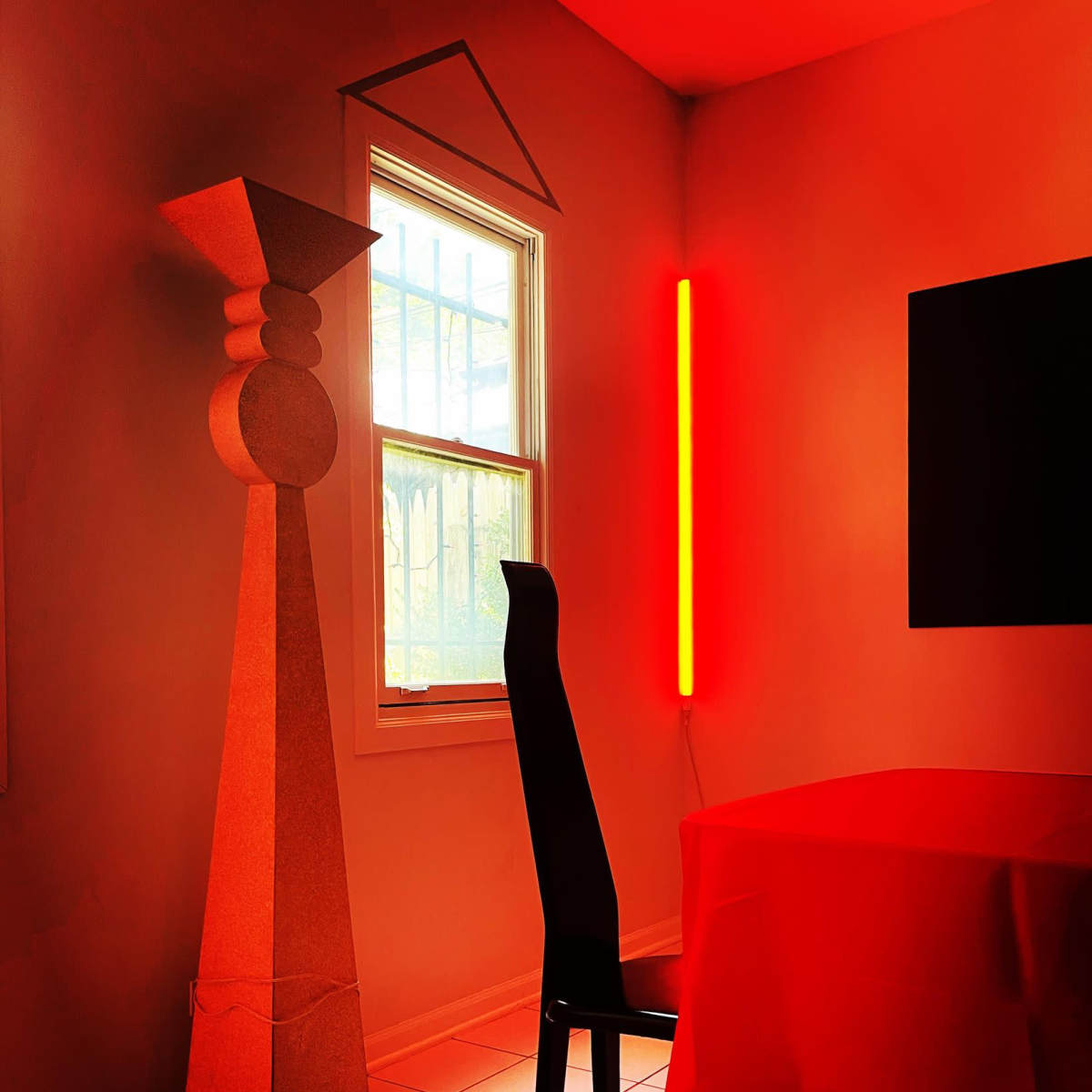 A room with a red tablecloth, a black chair, a tall sculptural element, and a neon red light illuminating the space near a window.