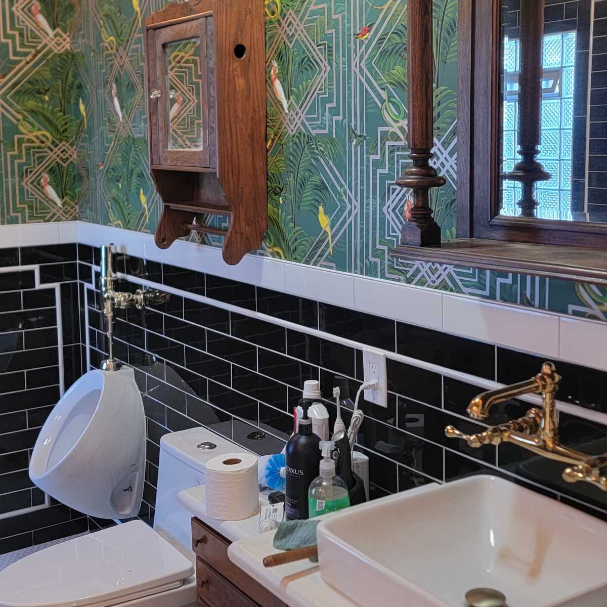 A bathroom featuring patterned green wallpaper, a wooden mirror frame, a black tiled wall, and a white toilet and sink.