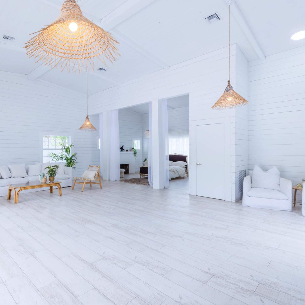 A bright, open room features white walls and wooden flooring, furnished with a sofa, chairs, and hanging woven light fixtures.