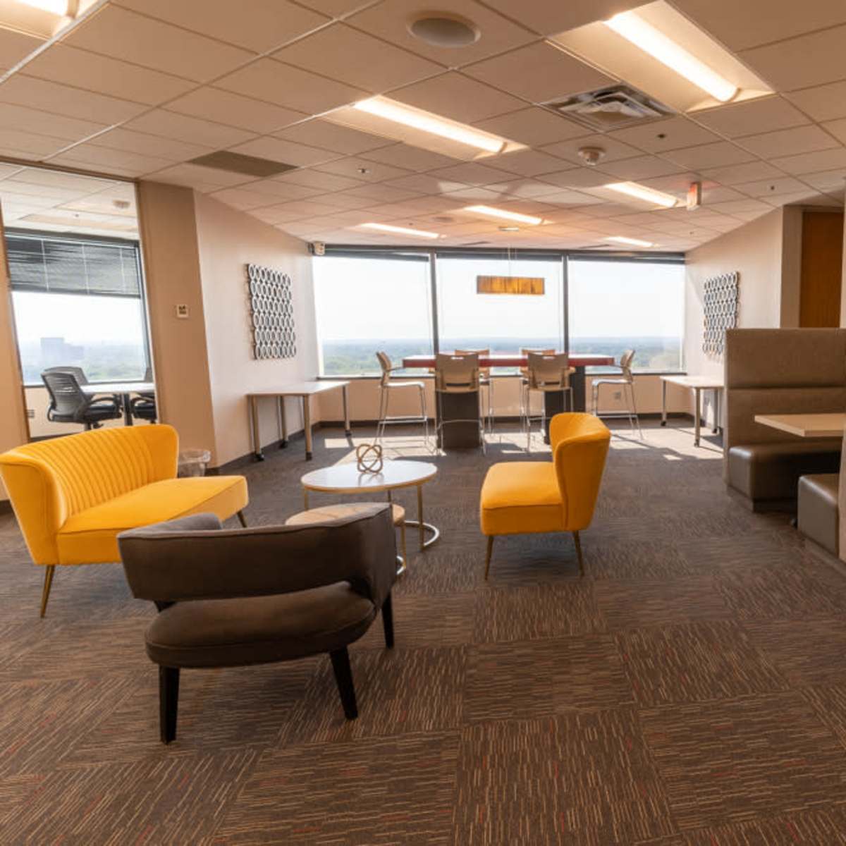 A modern office lounge area with yellow and gray furniture, small tables, and large windows overlooking a cityscape.