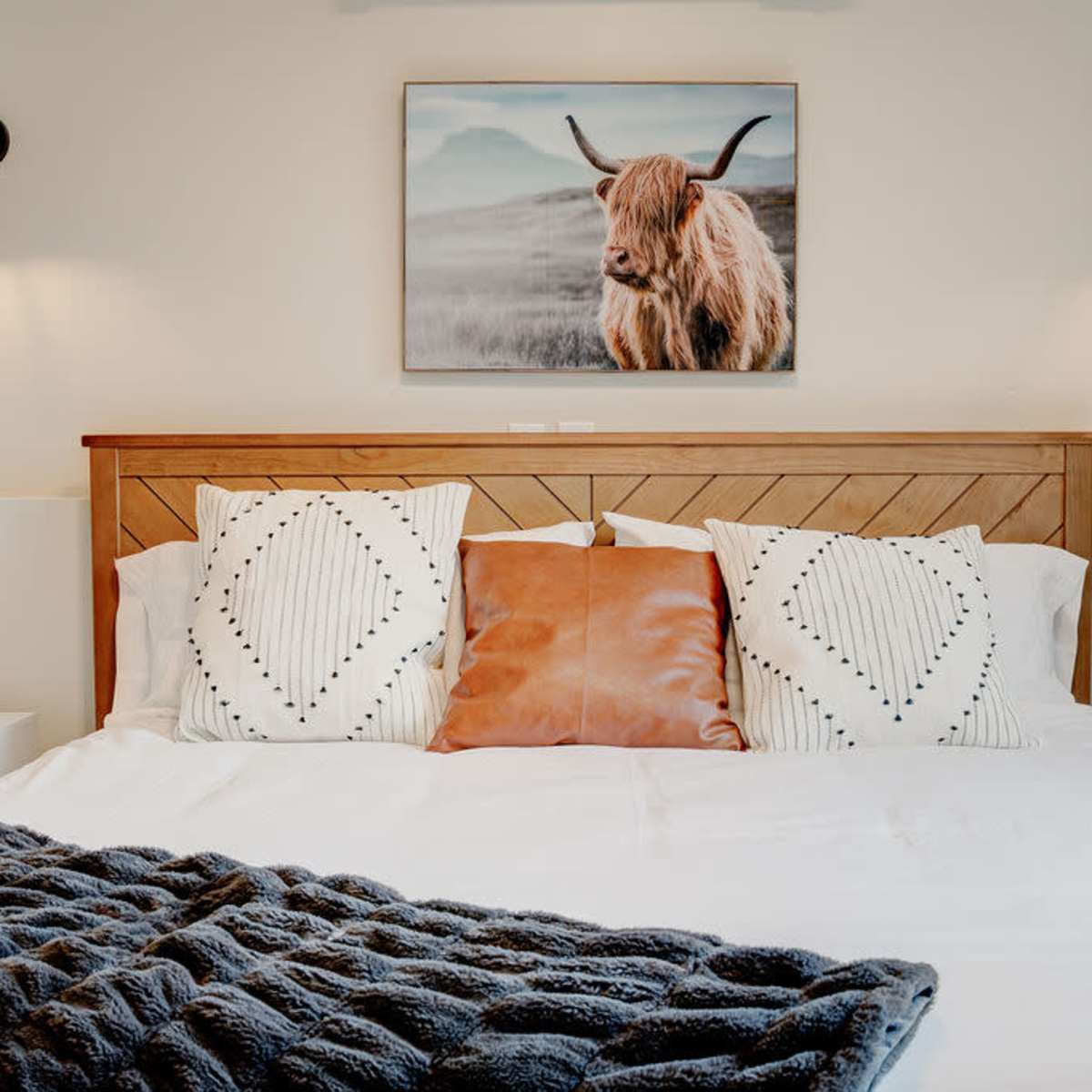 A neatly arranged bed with patterned pillows and a cozy blanket, adorned with a framed picture of a highland cow on the wall above.