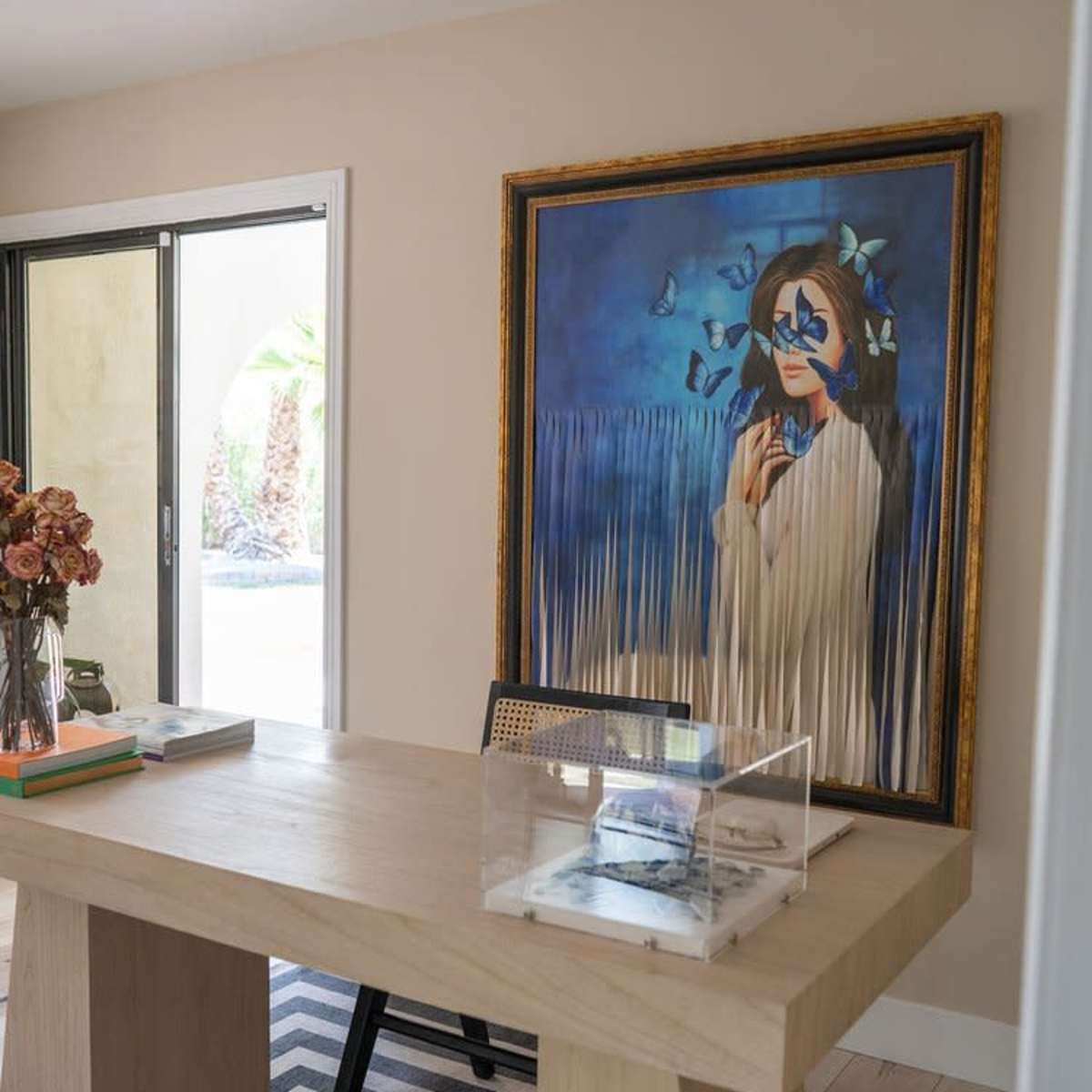A modern office space with a large wooden table, a framed painting of a woman with blue butterflies, and a striped rug underneath.