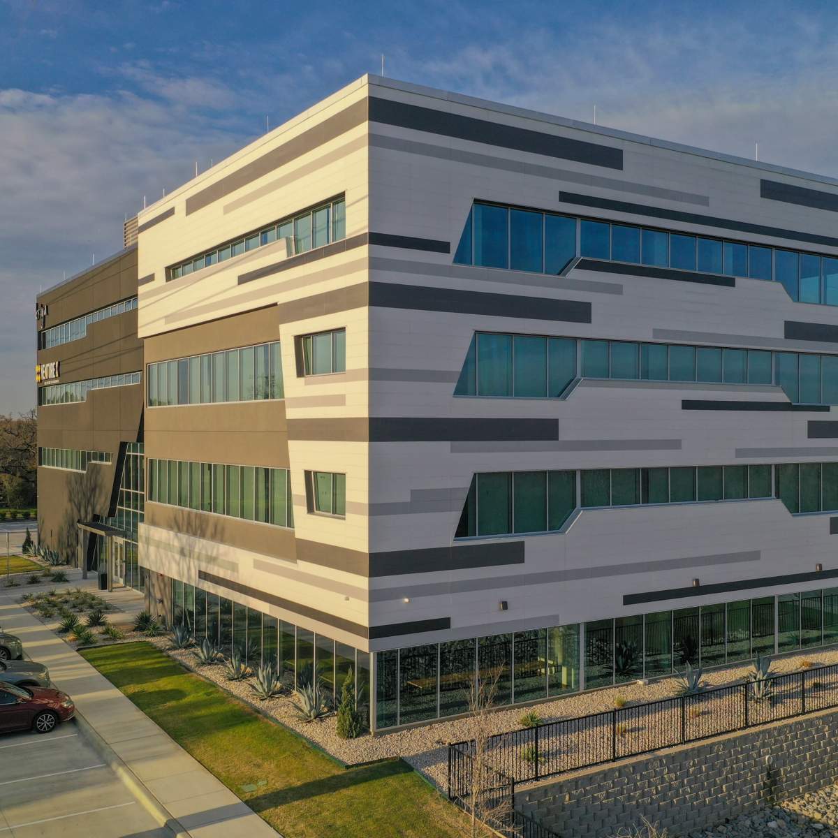 A modern, multi-story building with angular architectural features and large windows, set in a landscaped area with parked cars nearby.