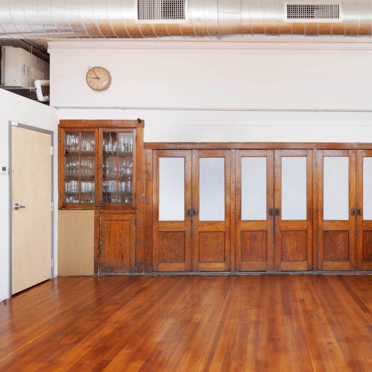 A spacious kitchen area featuring a wooden cabinet with glass doors, multiple wooden panel doors, and an adjacent modern kitchen counter with appliances against a backdrop of hardwood flooring.