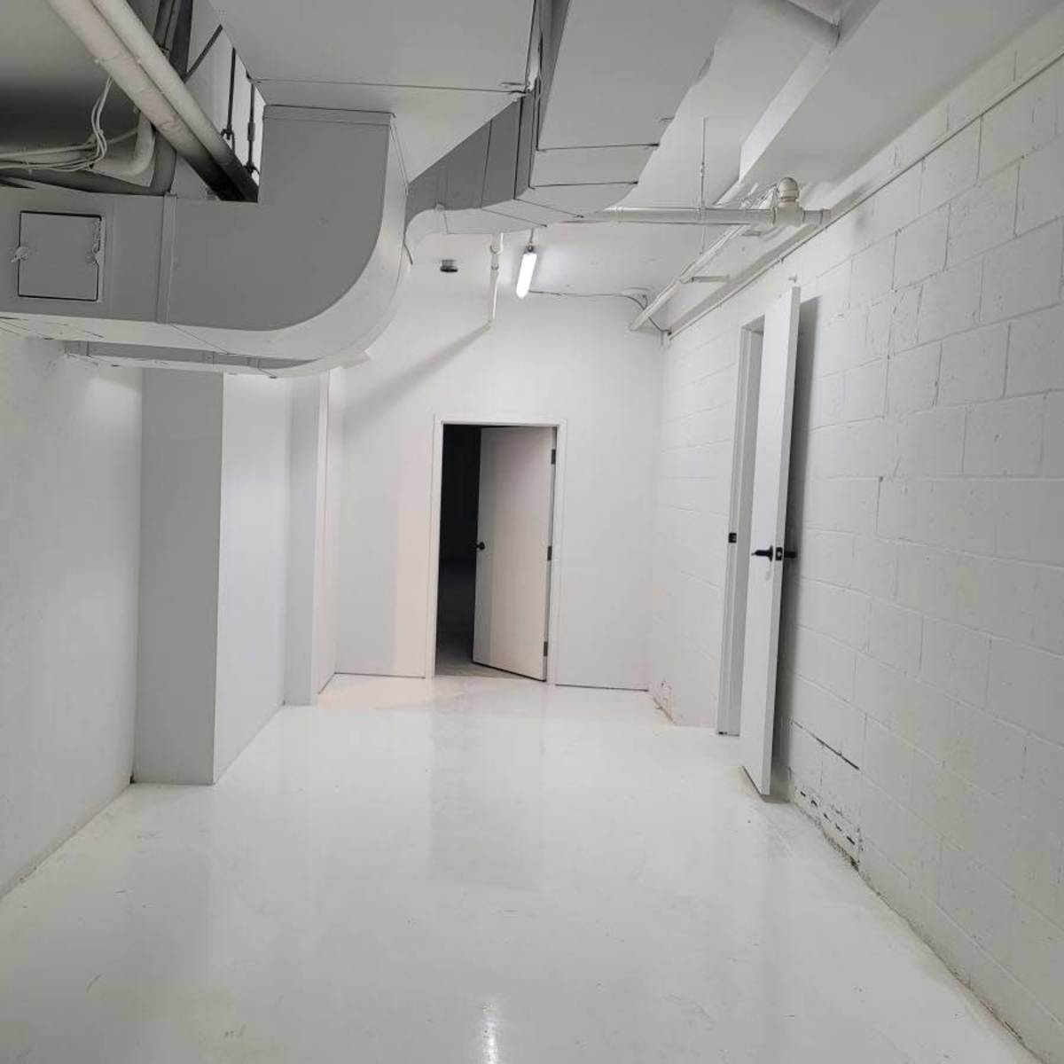 A sparsely furnished white corridor with two doors on the right and ventilation ducts above.