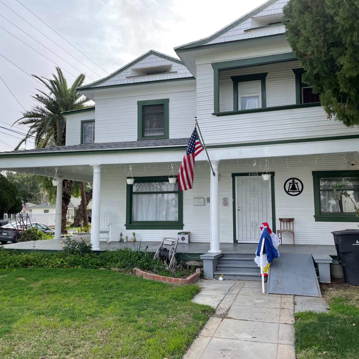 A two-story white house with green trim, an American flag hanging from the porch, and a well-maintained lawn.