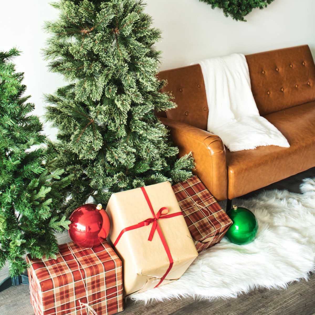 A decorated Christmas tree stands next to a cozy brown sofa, surrounded by wrapped gifts and holiday ornaments against a backdrop of a green wreath on the wall.